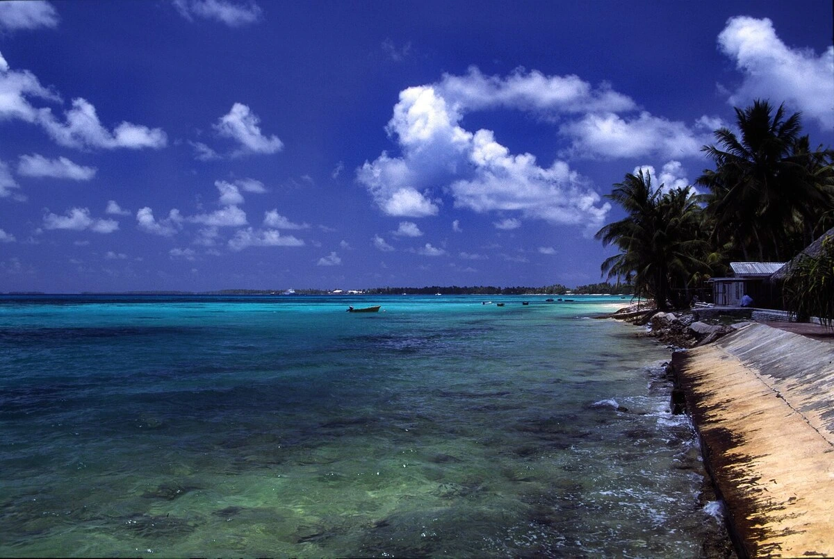 Playa de Funafuti en un día parcialmente soleado, con aguas cristalinas y vegetación tropical en la costa, típica de los paisajes insulares del Pacífico.