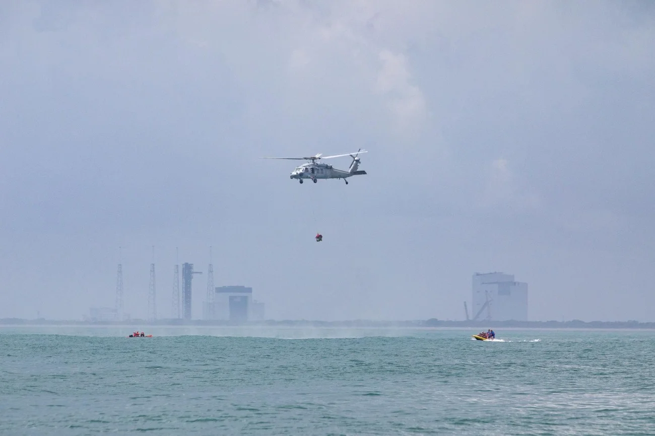 Helicóptero sobrevolando el mar con personal suspendido, instalaciones del Centro Espacial Kennedy visibles al fondo.