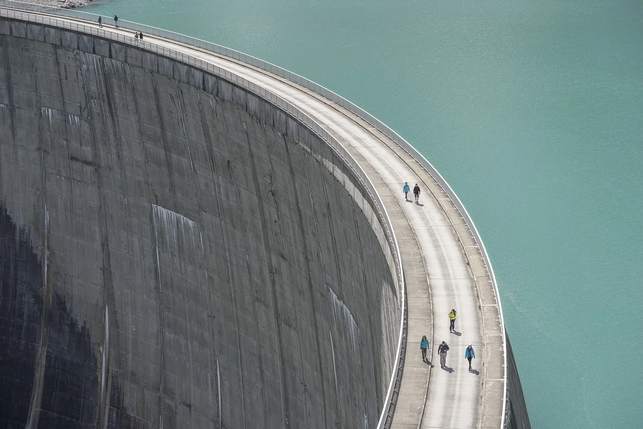 Personas caminando sobre la cresta de una represa alta con aguas turquesas de fondo