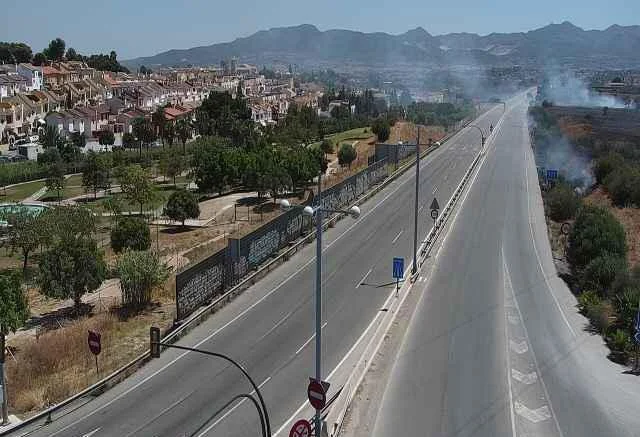 Vista aérea de una avenida en Málaga con humo por incendio en el margen derecho, cerca de zona residencial y montaña al fondo.