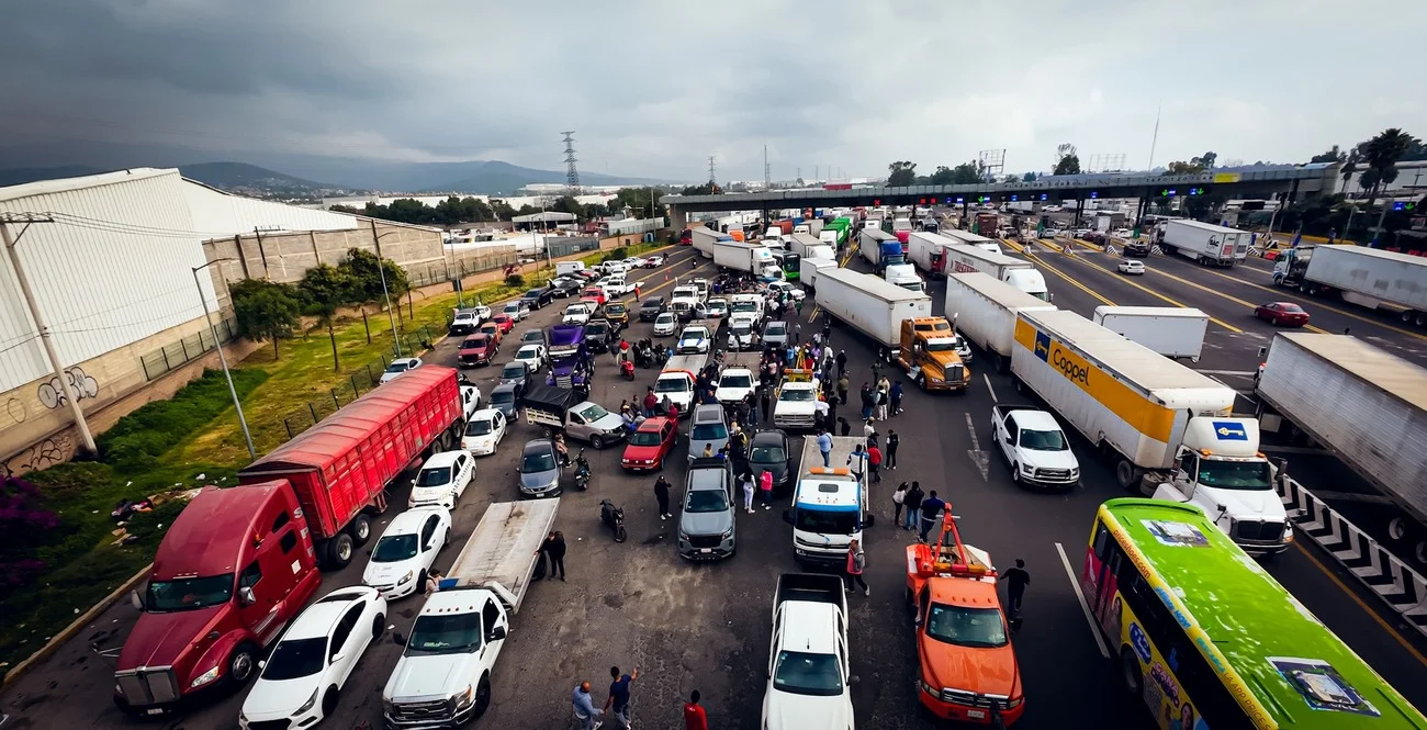 Panorámica de vehículos y personas bloqueando el acceso en una autopista de peaje bajo cielo nublado.
