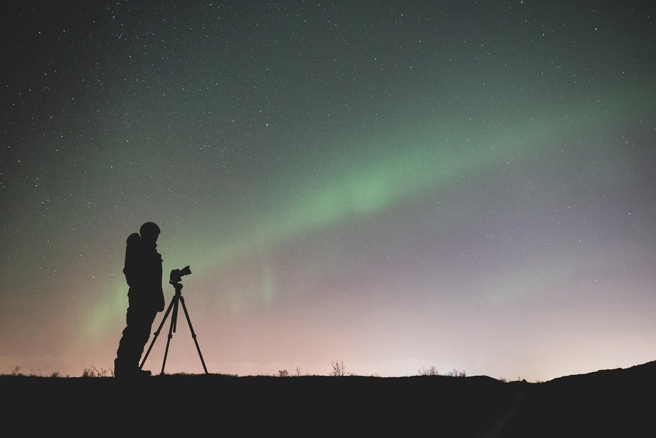 Persona observando el cielo estrellado y una aurora boreal con cámara y trípode en un paisaje nocturno