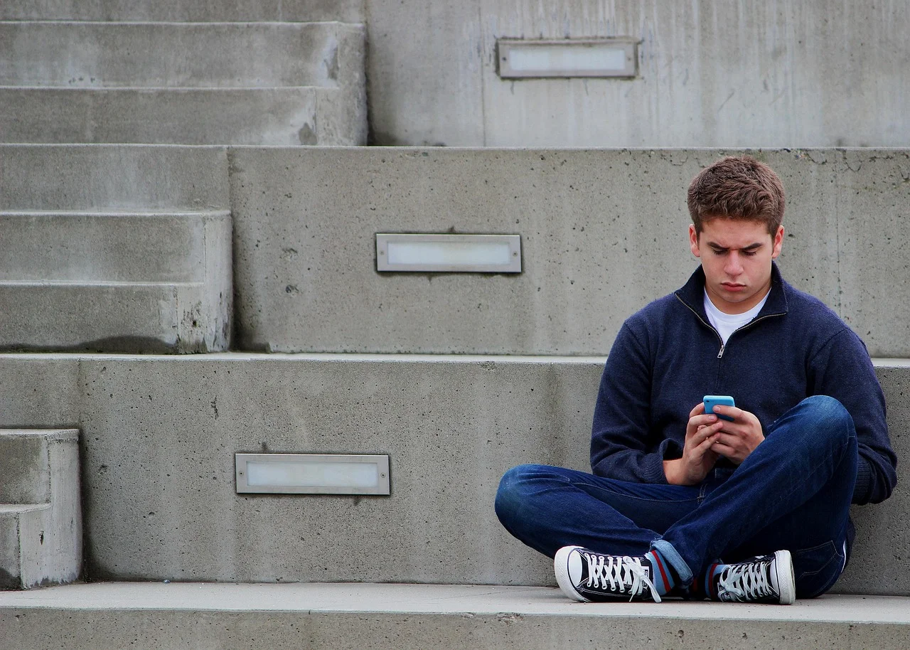 Adolescente sentado solo en unas escaleras mirando su teléfono móvil con gesto serio, representando la soledad juvenil