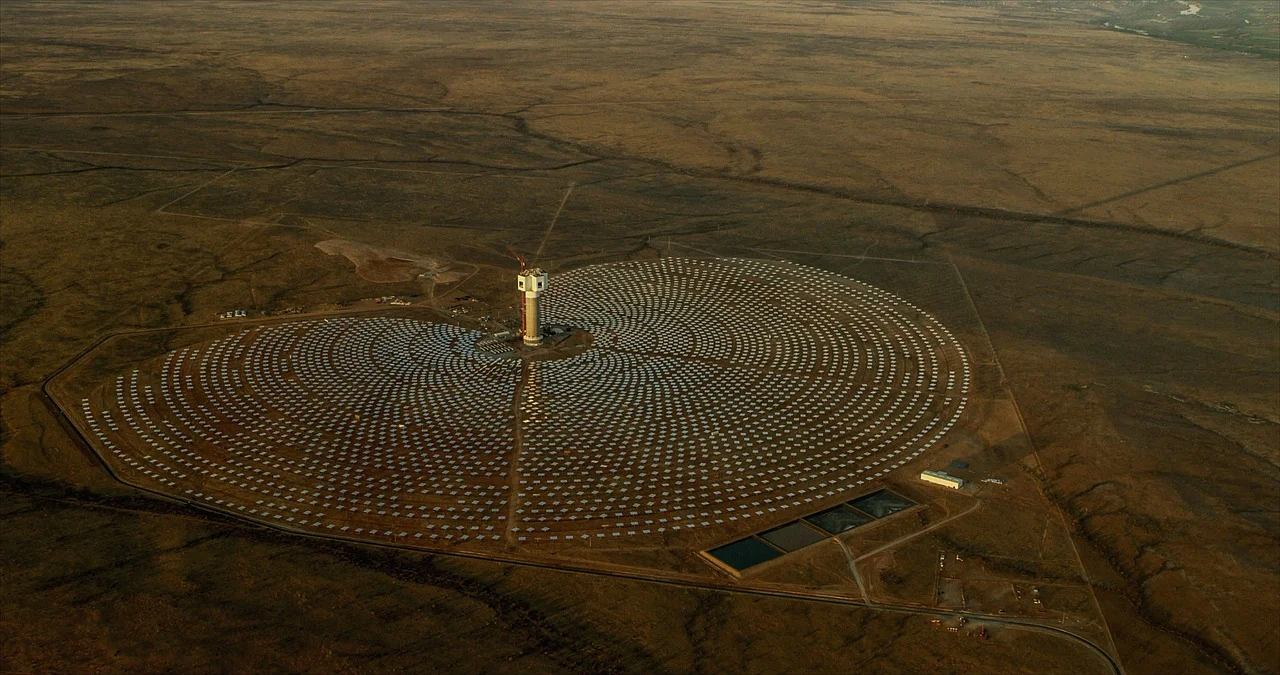 Planta termosolar con torre central y heliostatos brillando en el desierto, rodeada de montañas al fondo