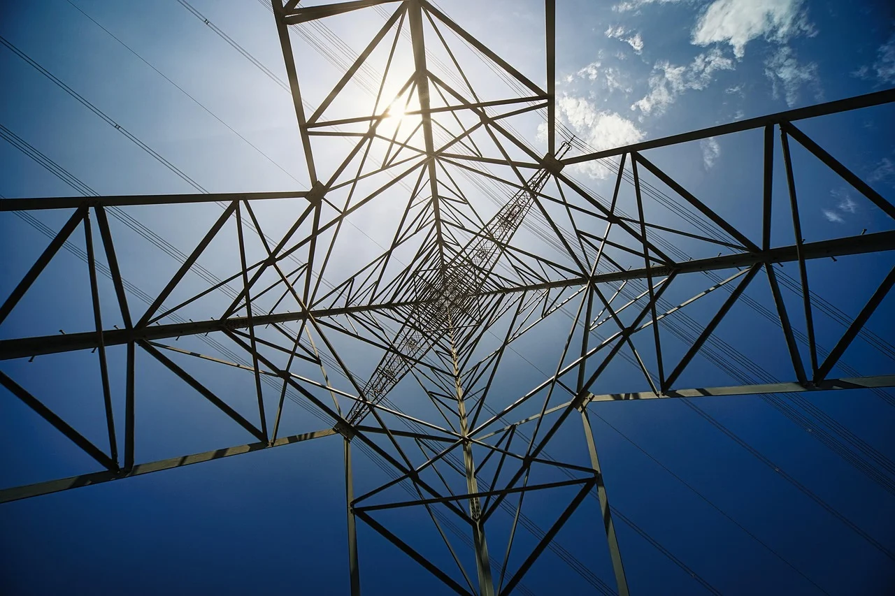 Estructura metálica de una torre de alta tensión vista desde abajo, con el cielo azul y el sol filtrándose entre los cables eléctricos.