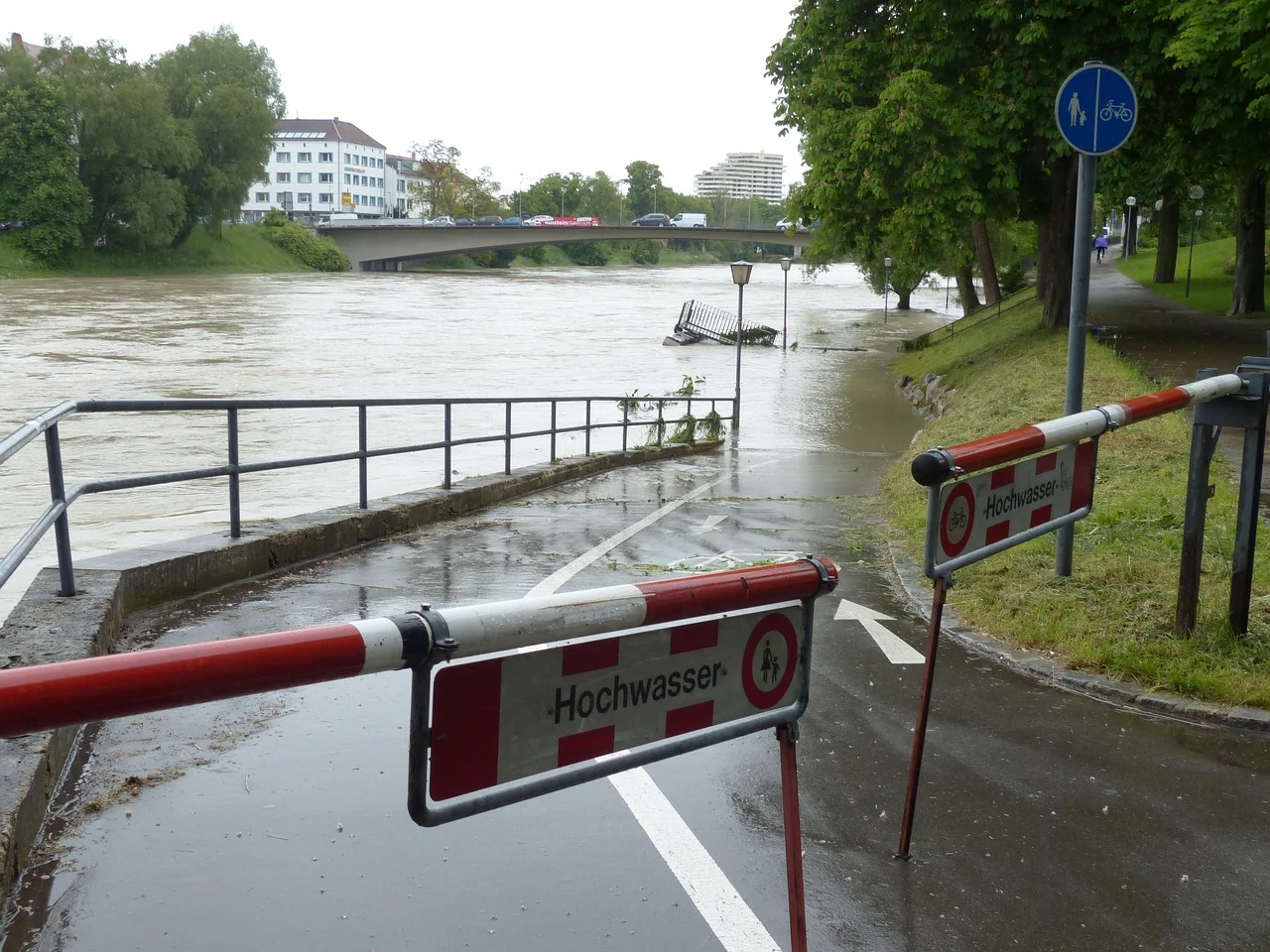 Inundación del río con paso peatonal anegado y señal de “Hochwasser” cerrando el acceso