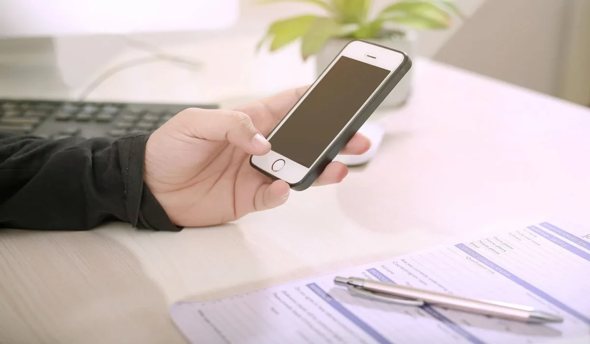 Persona sostiene un teléfono inteligente frente al ordenador mientras trabaja en un escritorio.