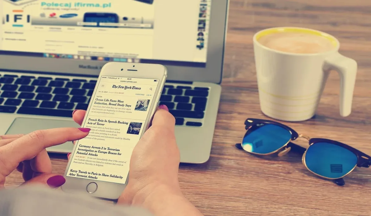 Persona usando su teléfono móvil mientras lee noticias en línea, con un portátil y una taza de café al fondo.