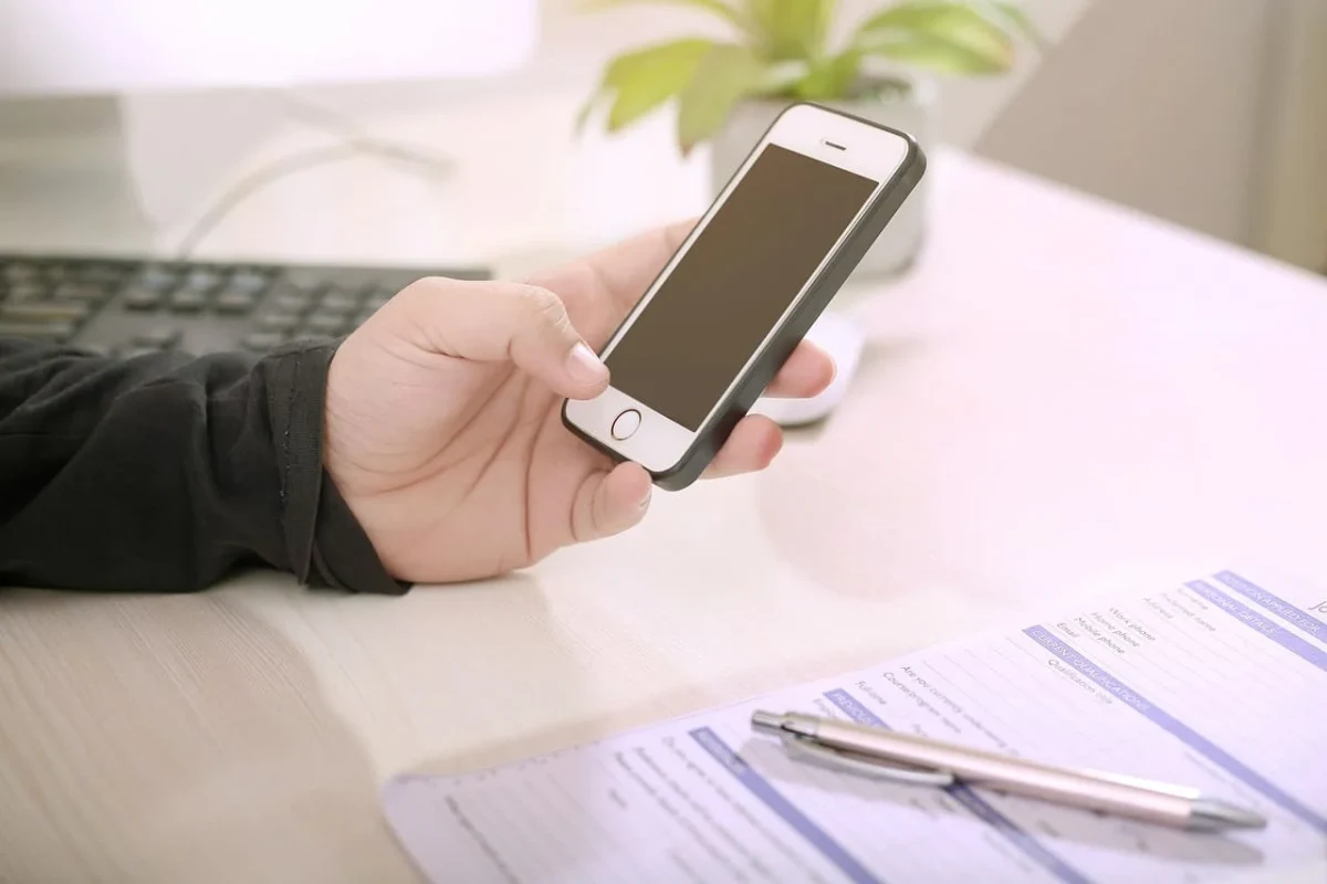Persona sostiene un teléfono inteligente frente al ordenador mientras trabaja en un escritorio.