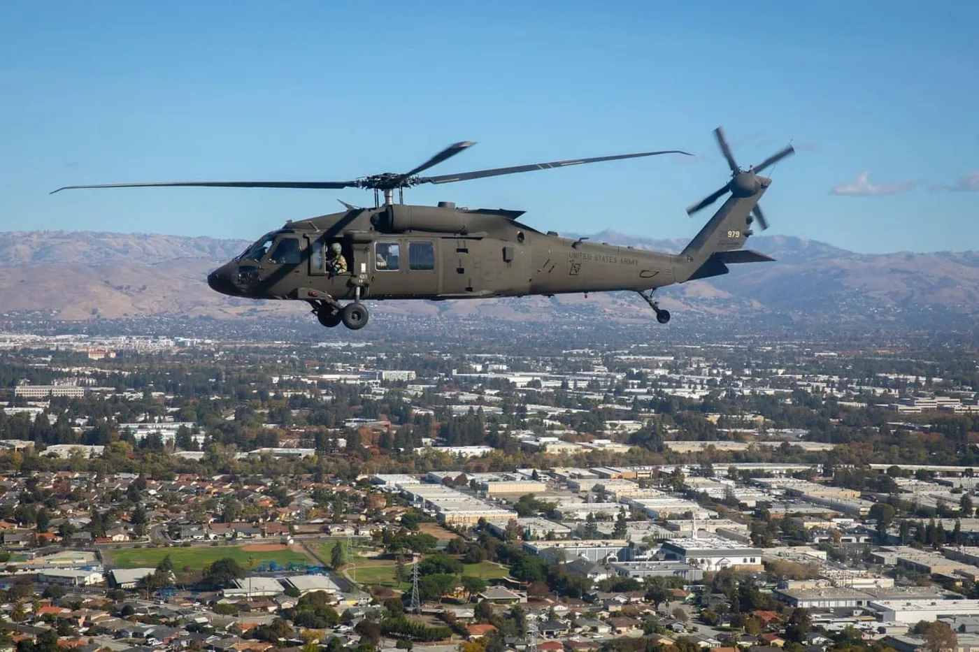 Helicóptero militar UH-60 Black Hawk del Ejército de los Estados Unidos volando sobre una ciudad con montañas al fondo.