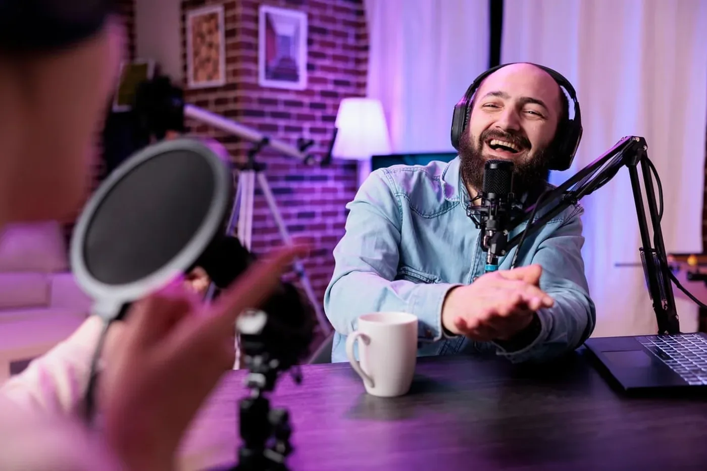 Hombre con auriculares grabando un pódcast frente a un micrófono y una laptop en estudio informal.