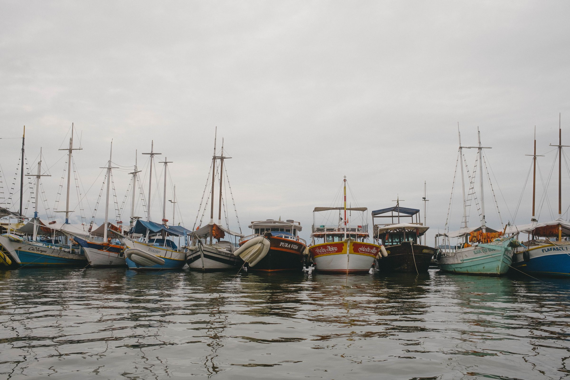 A fleet of colorful fishing boats and sailboats with tall masts are moored in calm harbor waters under an overcast sky, their reflections shimmering on the water’s surface