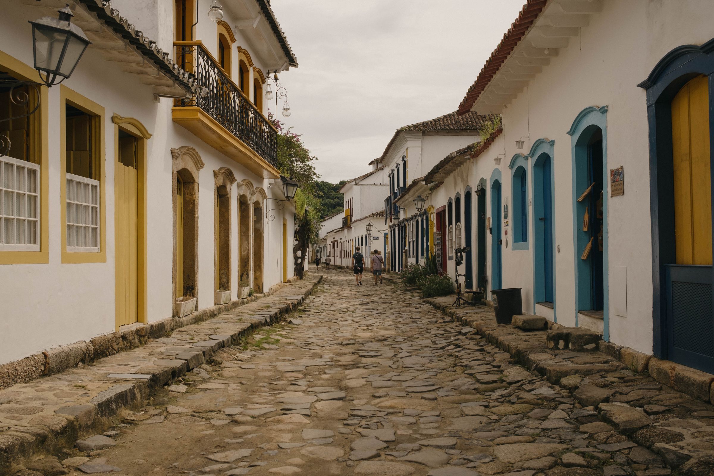 A cobblestone street lined with white colonial buildings featuring colorful window and door frames in yellow and turquoise stretches into the distance, with a few pedestrians walking along the historic stone-paved road.