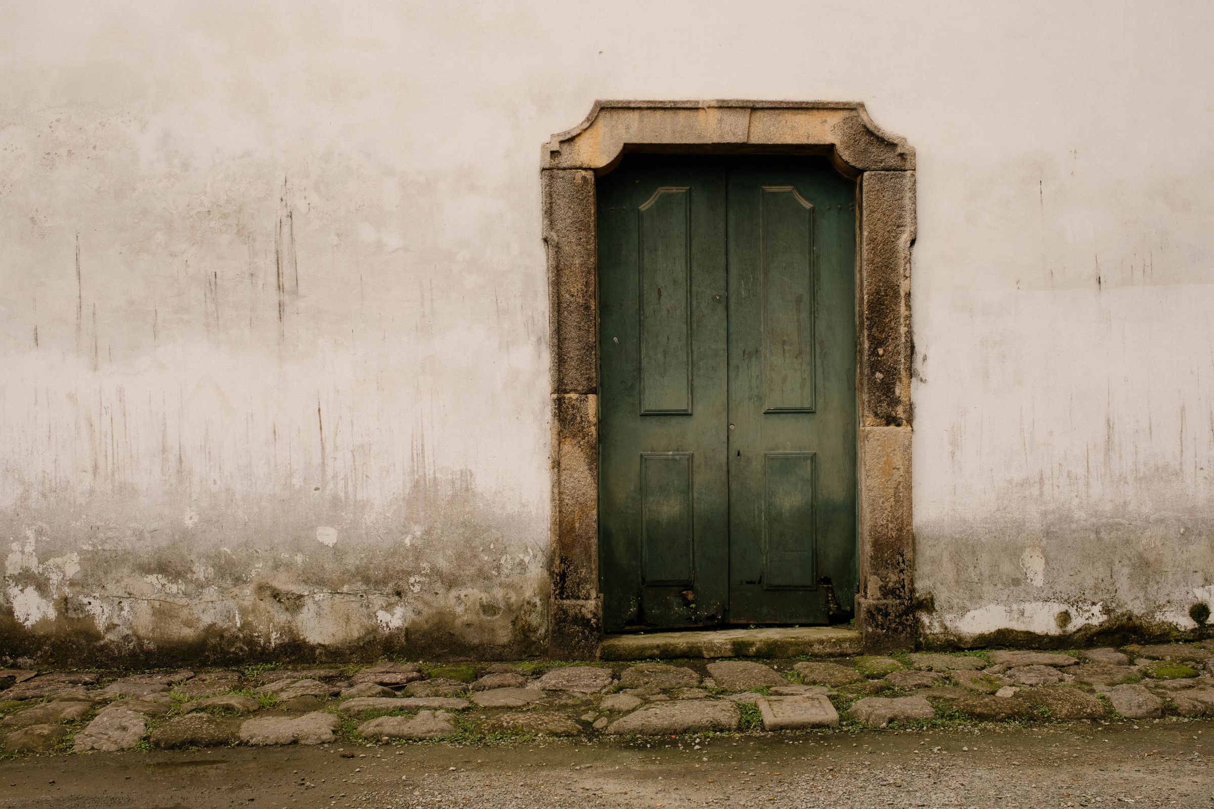 A weathered green wooden door with carved stone frame stands centered on a white colonial wall, with moss growing between the cobblestones at its base and age-darkened stains marking the lower portion of the facade.
