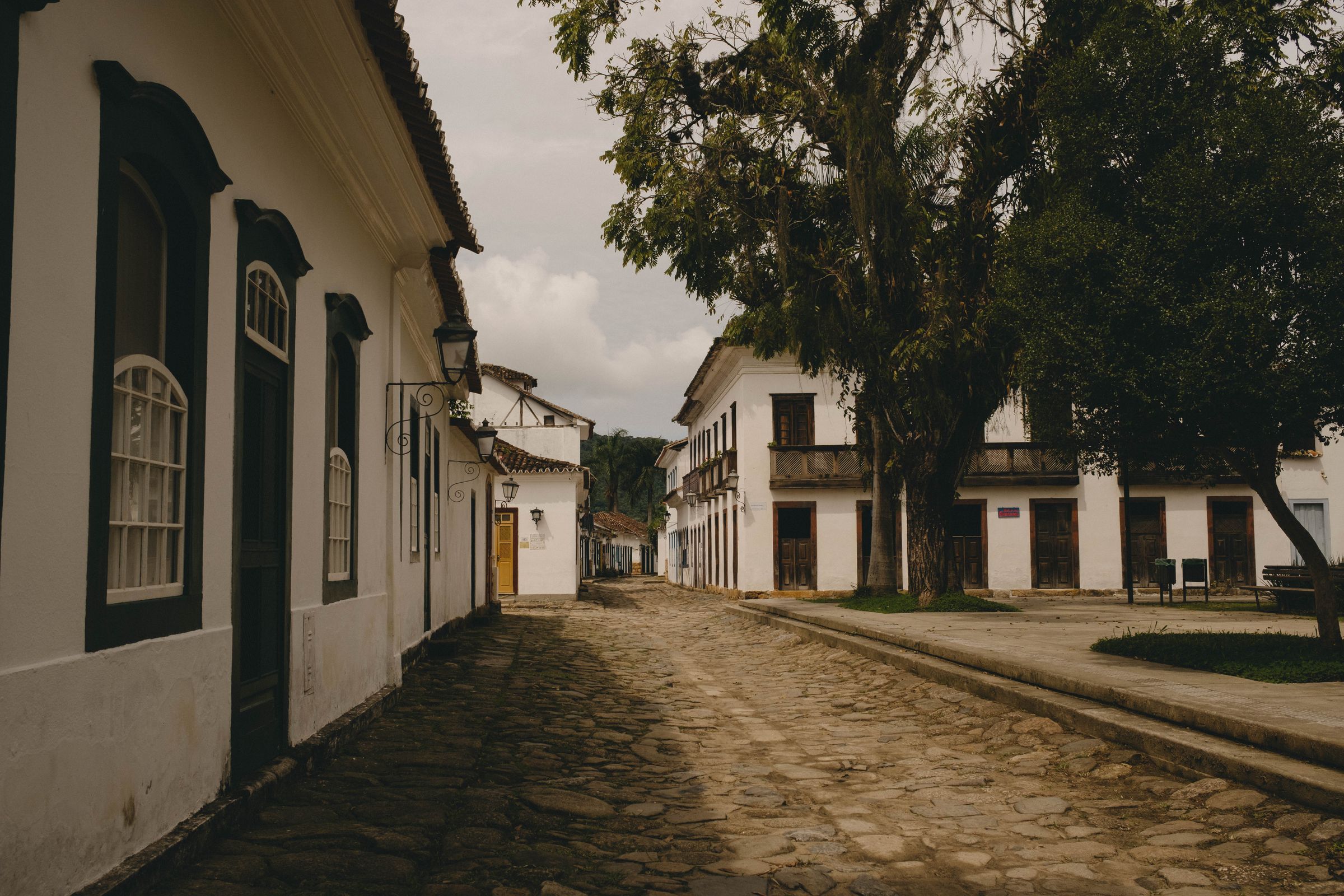 A cobblestone street curves past white colonial buildings with dark green shutters and trim, while a large tree casts shadows over the historic town square under an overcast sky.