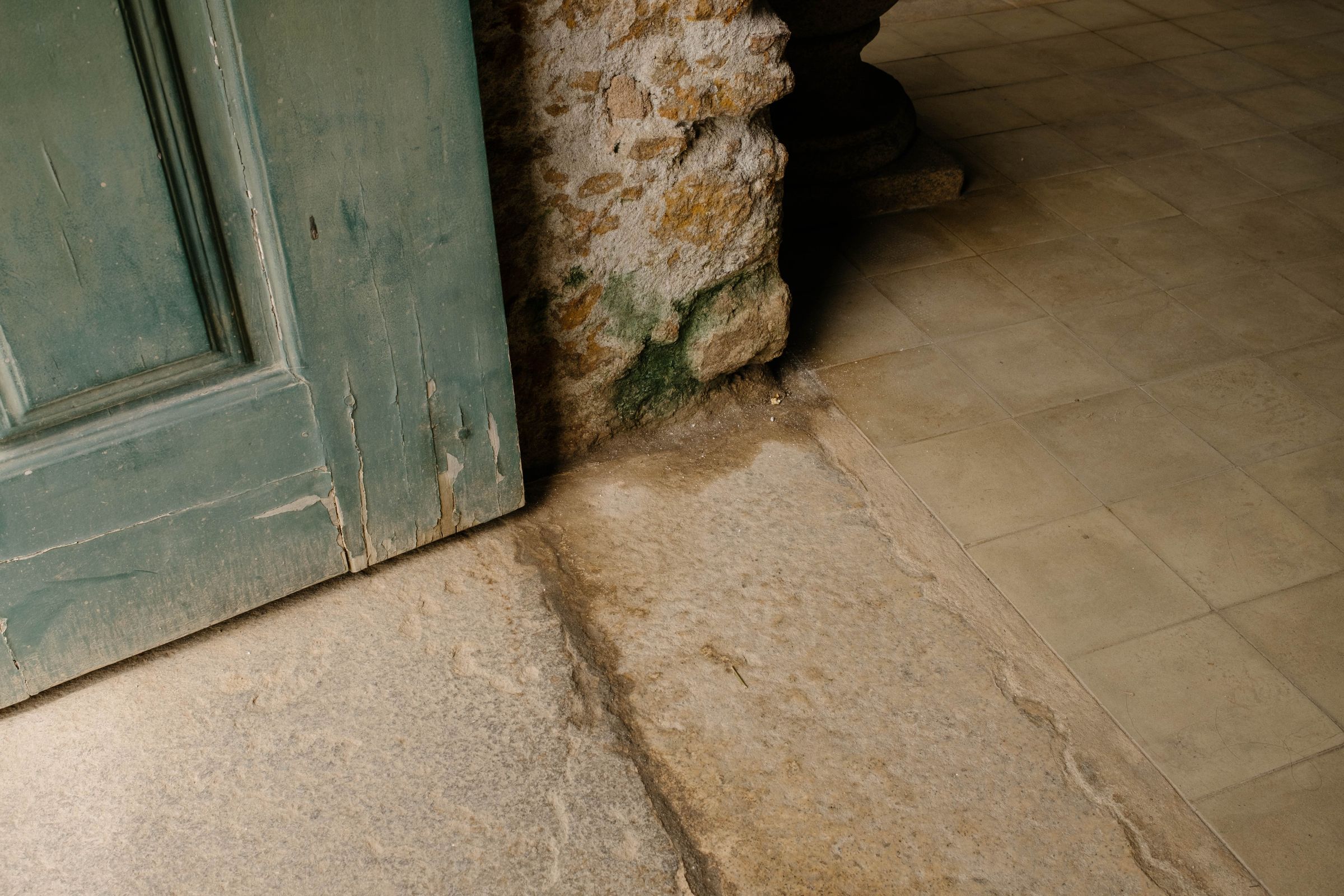 A weathered turquoise door frame meets moss-covered stone walls at the base, with worn stone tiles creating geometric patterns on the ground and showing centuries of age and wear.