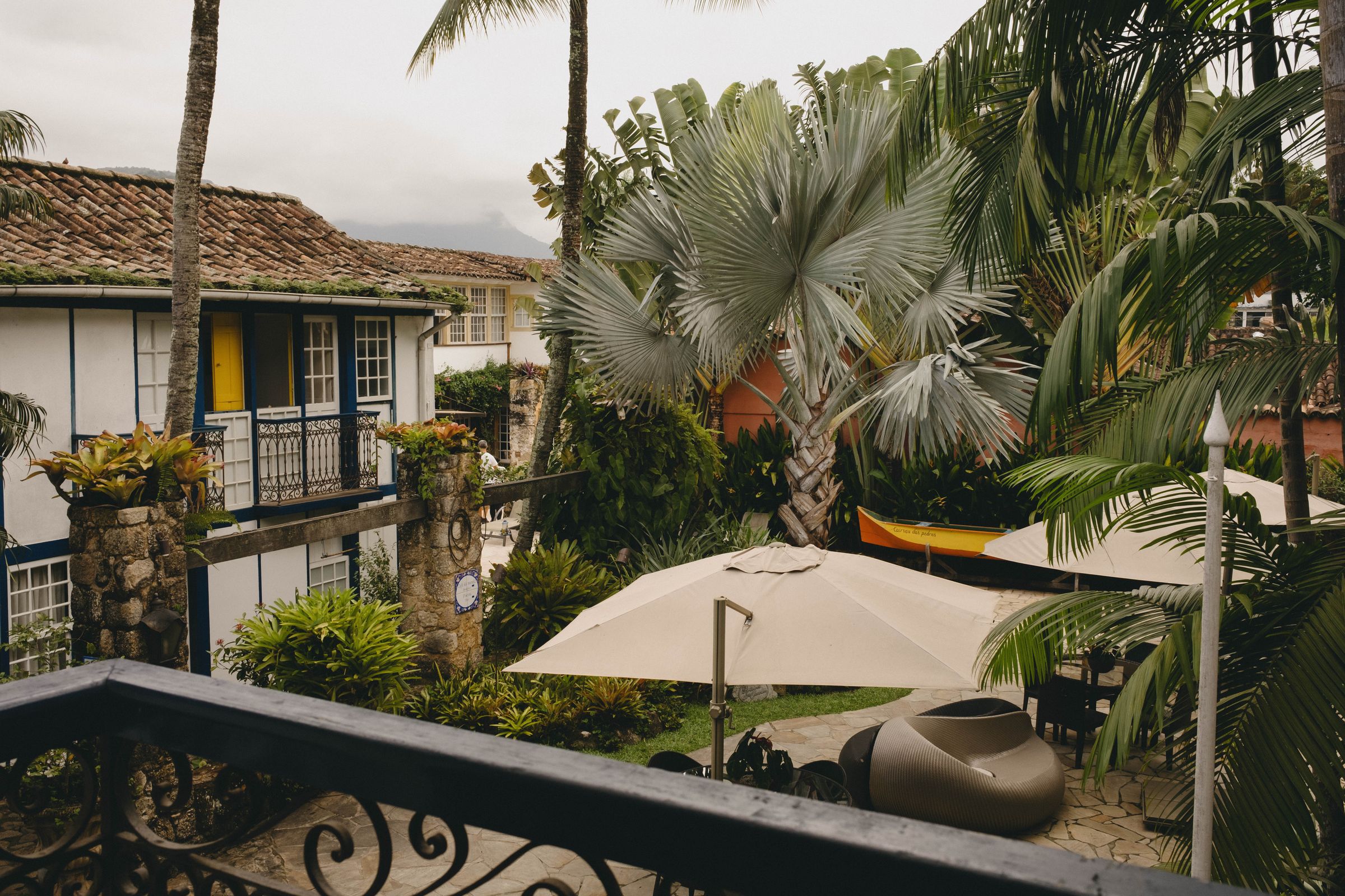 A tropical courtyard scene framed by ornate iron railings showcases colonial architecture with terracotta tile roofs and colorful shutters nestled among lush palm trees and tropical vegetation. The composition captures the layered textures of the scene, from the decorative ironwork in the foreground to the white patio umbrella and dense foliage that create a sense of depth and tropical abundance.
