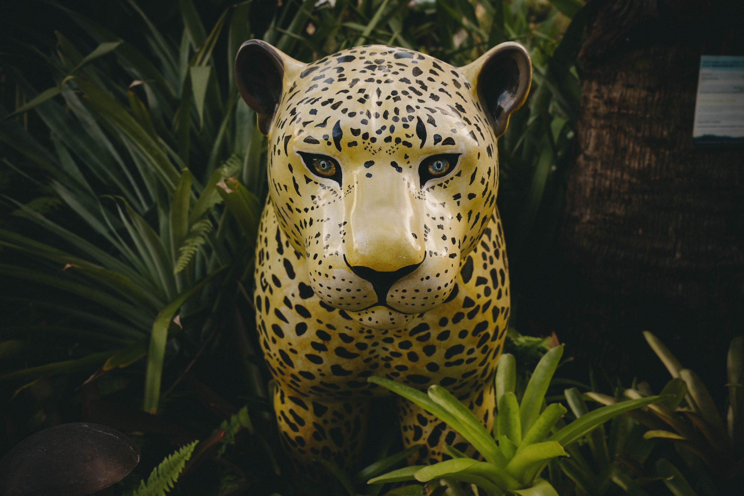A vibrant ceramic jaguar sculpture with piercing blue eyes and meticulously painted spotted markings emerges from dense tropical foliage, its golden coat creating a striking contrast against the dark green vegetation. The piece captures the intense, watchful gaze of the big cat while nestled among palm fronds and jungle plants, blending artistry with the wild aesthetic of its natural habitat.