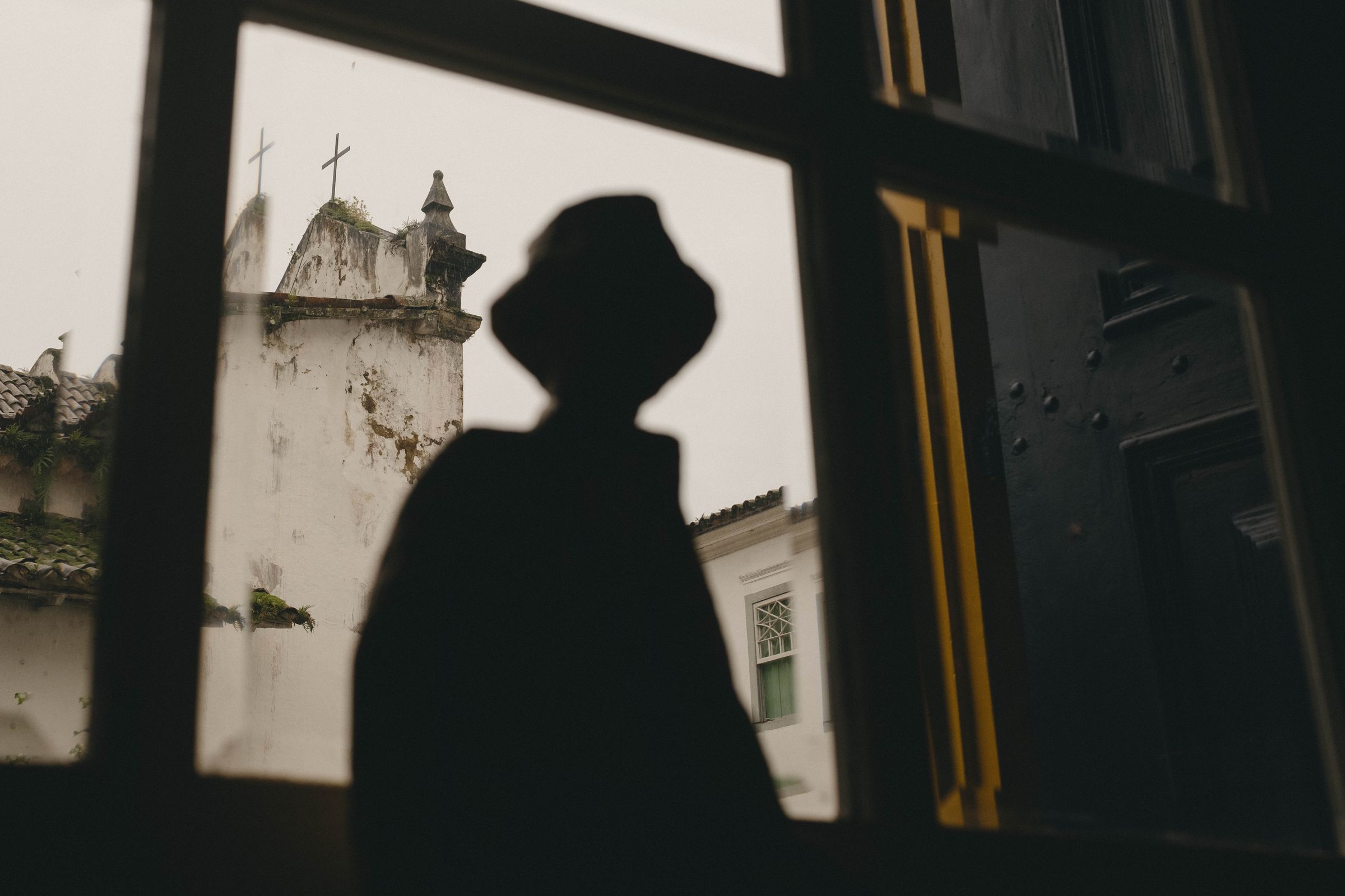 A dramatic silhouette of a statue of a person wearing a hat is cast against a white colonial wall, with a weathered church bell tower topped by crosses visible in the background. The composition creates a compelling layered narrative, juxtaposing the anonymous human shadow in the foreground with the enduring religious architecture that speaks to centuries of history.
