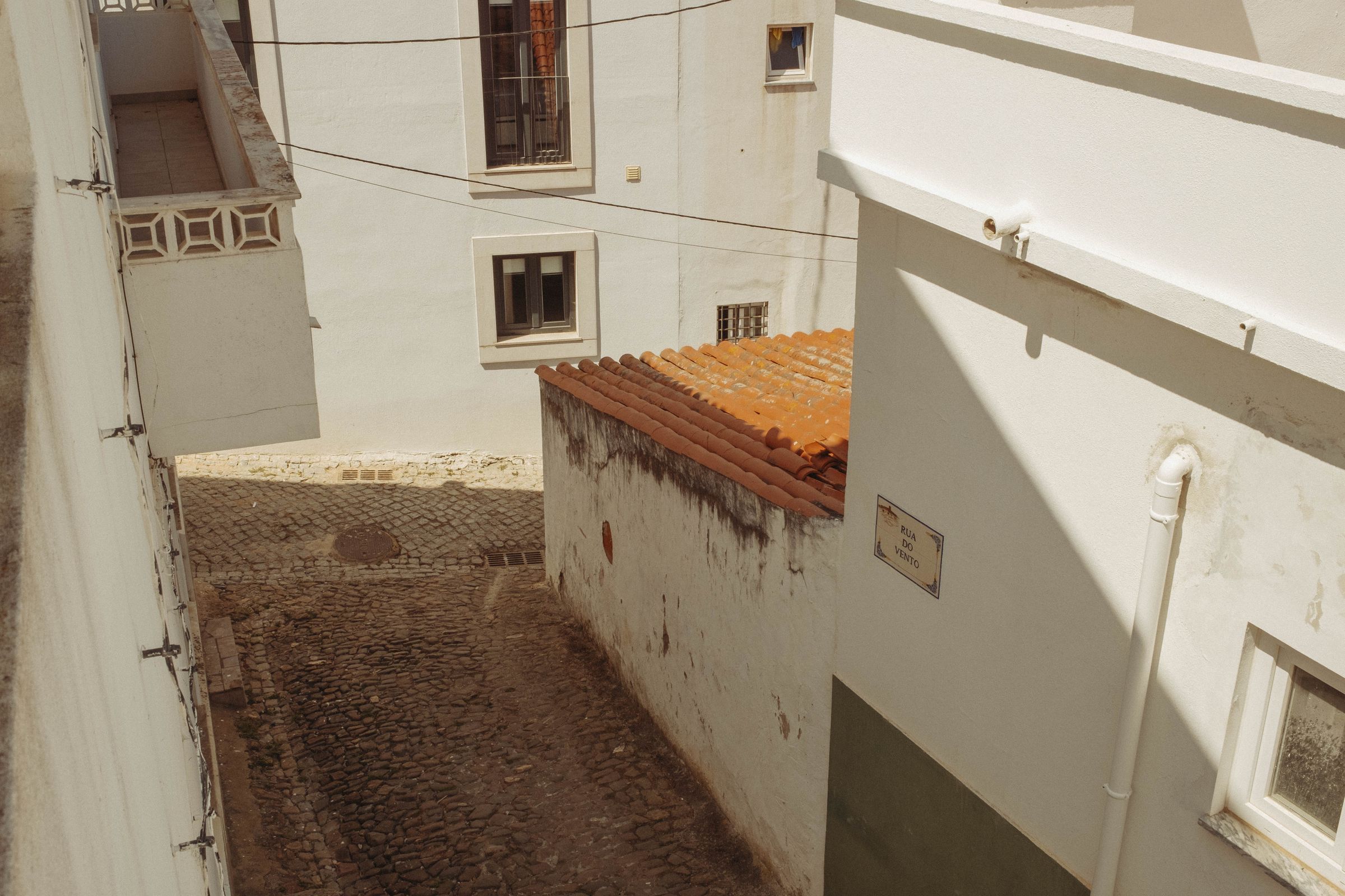 A narrow cobblestone alley winds between whitewashed buildings with weathered walls and small windows, descending toward an archway below. A terracotta-roofed addition juts into the passage, its aged surface contrasting with the bright white walls that catch harsh shadows in the afternoon light.