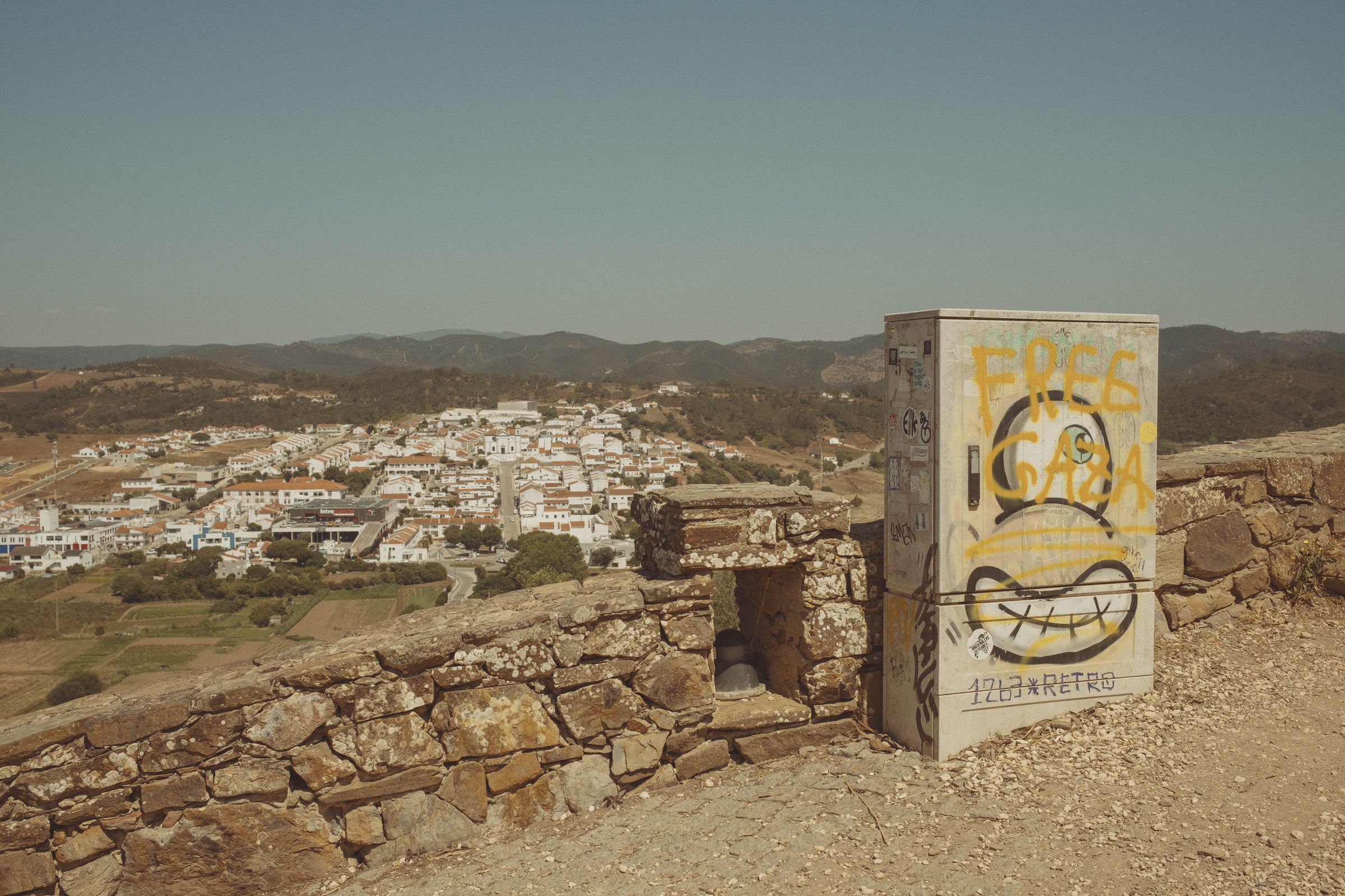 A weathered utility box painted with yellow graffiti reading 'FREE GAZA' stands on castle ruins overlooking the whitewashed town spread across hillsides below. The ancient stone walls frame the modern political statement against a landscape of terracotta roofs, rolling hills, and distant mountains under a hazy sky.