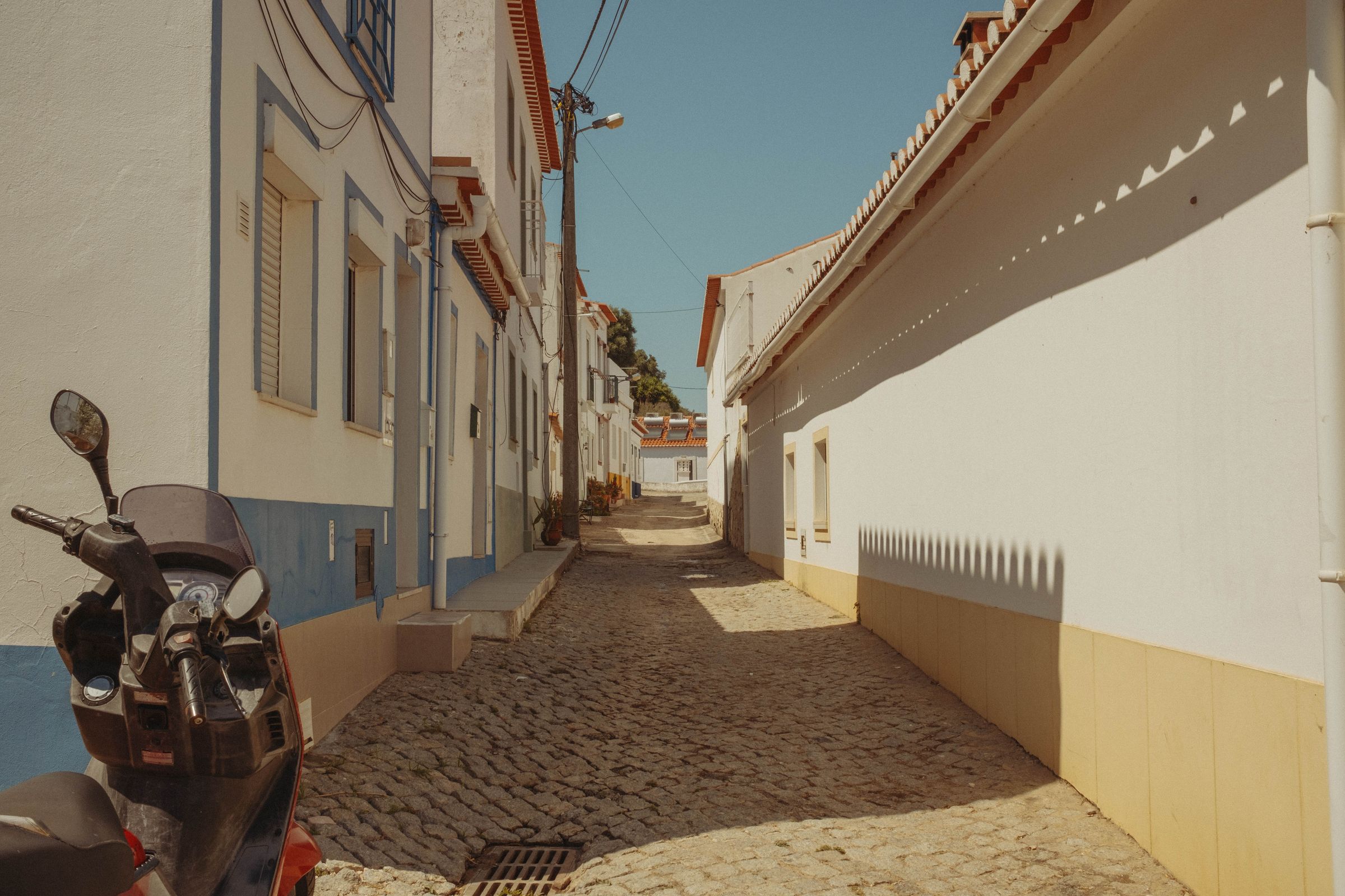 A narrow cobblestone alley climbs between whitewashed buildings trimmed in blue and yellow, their terracotta roofs creating a rhythmic pattern against the clear sky. A parked scooter rests on the left while power lines cross overhead, and the steep lane leads upward toward more white houses in the bright afternoon sun.