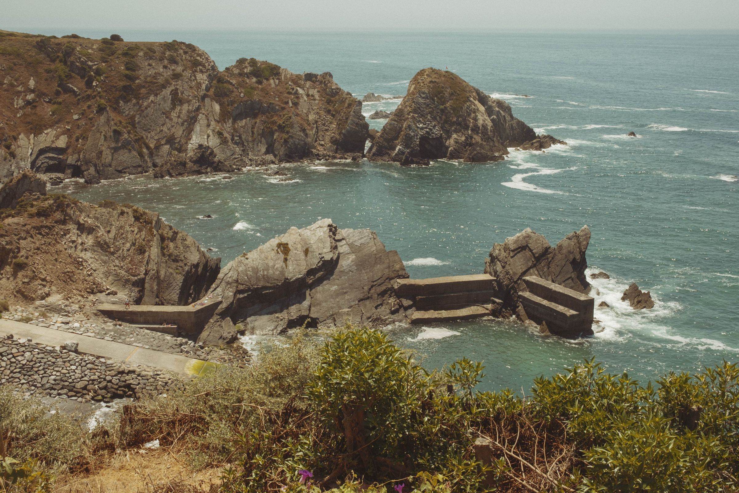 Dramatic rock formations jut from turquoise waters along the rugged Alentejo coast, with waves breaking around jagged outcrops and sea stacks. Moss-covered vegetation clings to the clifftop in the foreground, while eroded rocks reveal distinct geological layers where the wild Atlantic meets the land.