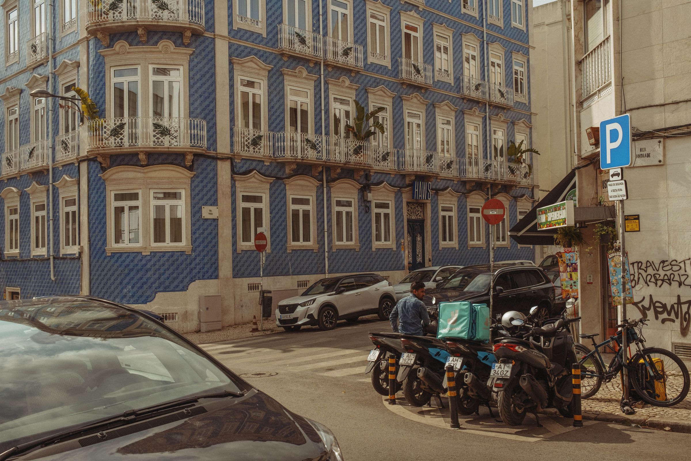 A delivery rider on a scooter pauses on a Lisbon street in front of a striking blue azulejo-tiled building with ornate white window frames and wrought-iron balconies. The traditional Portuguese facade, covered in diamond-patterned ceramic tiles, contrasts with the modern scene of parked cars, motorcycles, and a turquoise delivery bag.