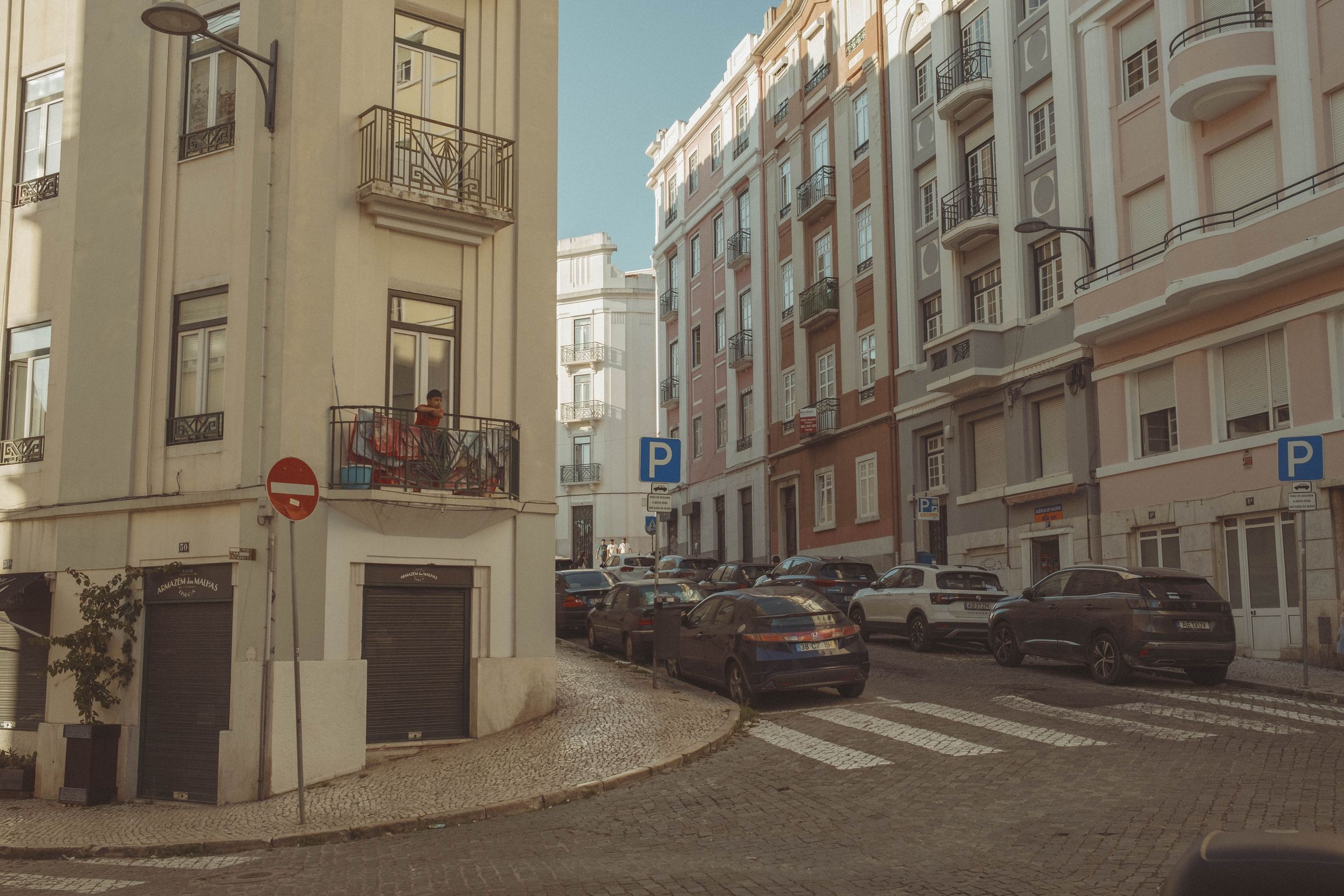 A person relaxes on a balcony overlooking a cobblestone street lined with parked cars and pastel apartment buildings in shades of cream, pink, and beige. The quiet residential block features ornate iron railings, Art Deco details, and parking signs, with a figure visible in the distance walking up the gently curving lane.