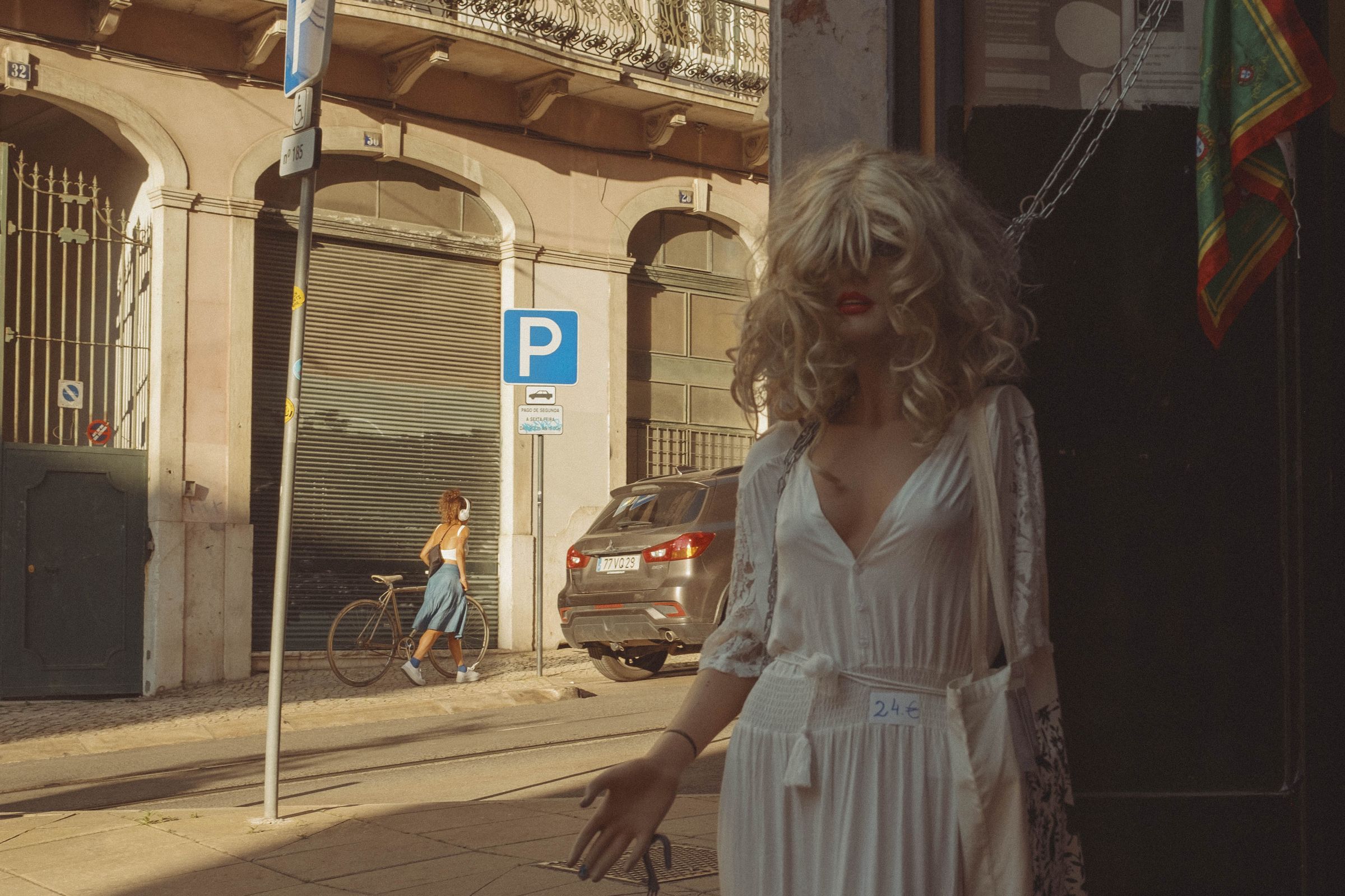 A mannequin of a woman in a white dress walks down a sunny street past arched storefronts with shuttered shops, while another figure pushes a bicycle in the background. Golden afternoon light illuminates the beige building facade with its ornate iron balcony and blue parking sign, creating long shadows across the pavement.