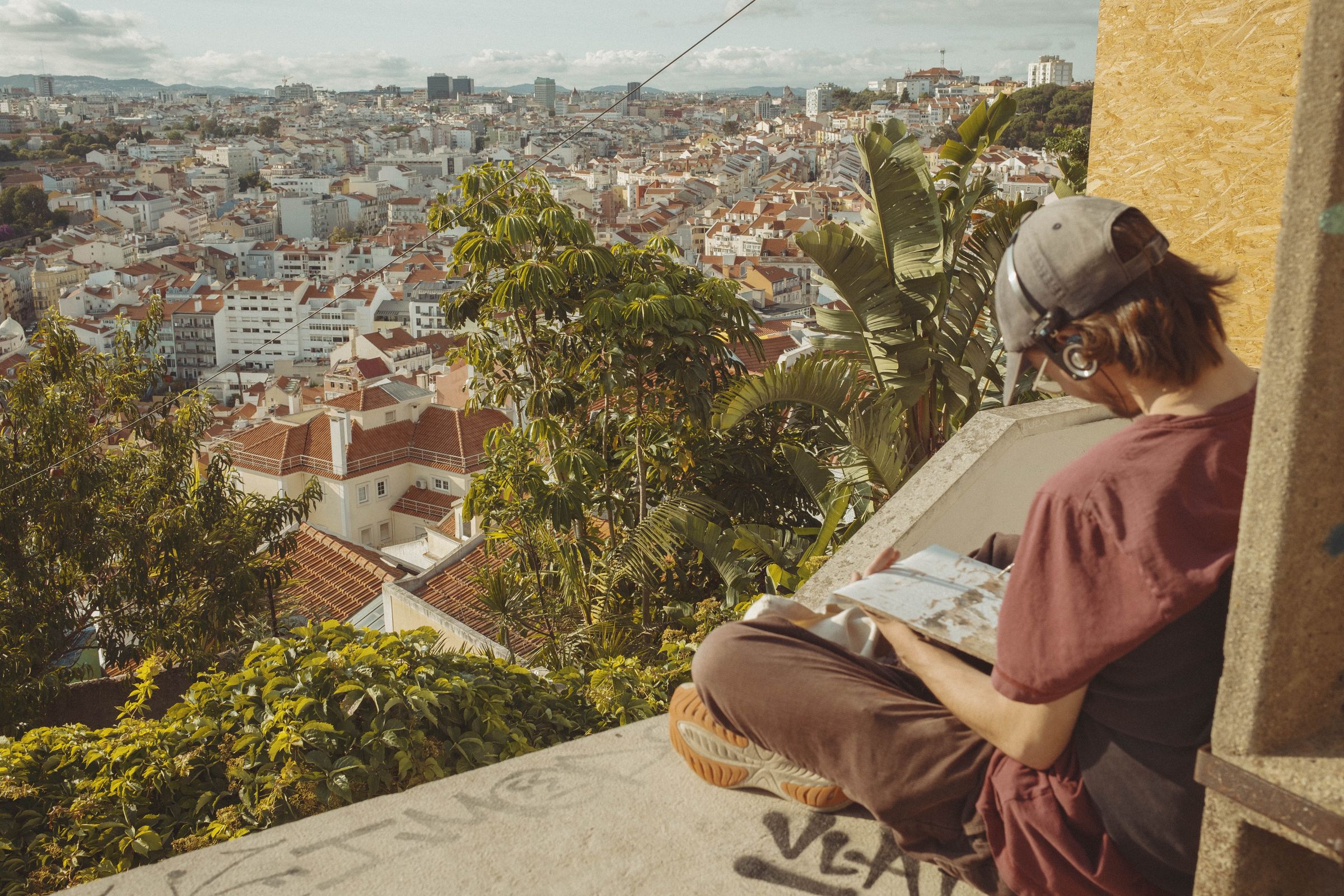 A person in a baseball cap and rose-colored clothing sits on a stone ledge sketching what they see in a book, with Lisbon’s vast cityscape of terracotta roofs spreading below. Lush tropical plants, including banana leaves, frame the foreground while distant hills rise under a cloudy sky.