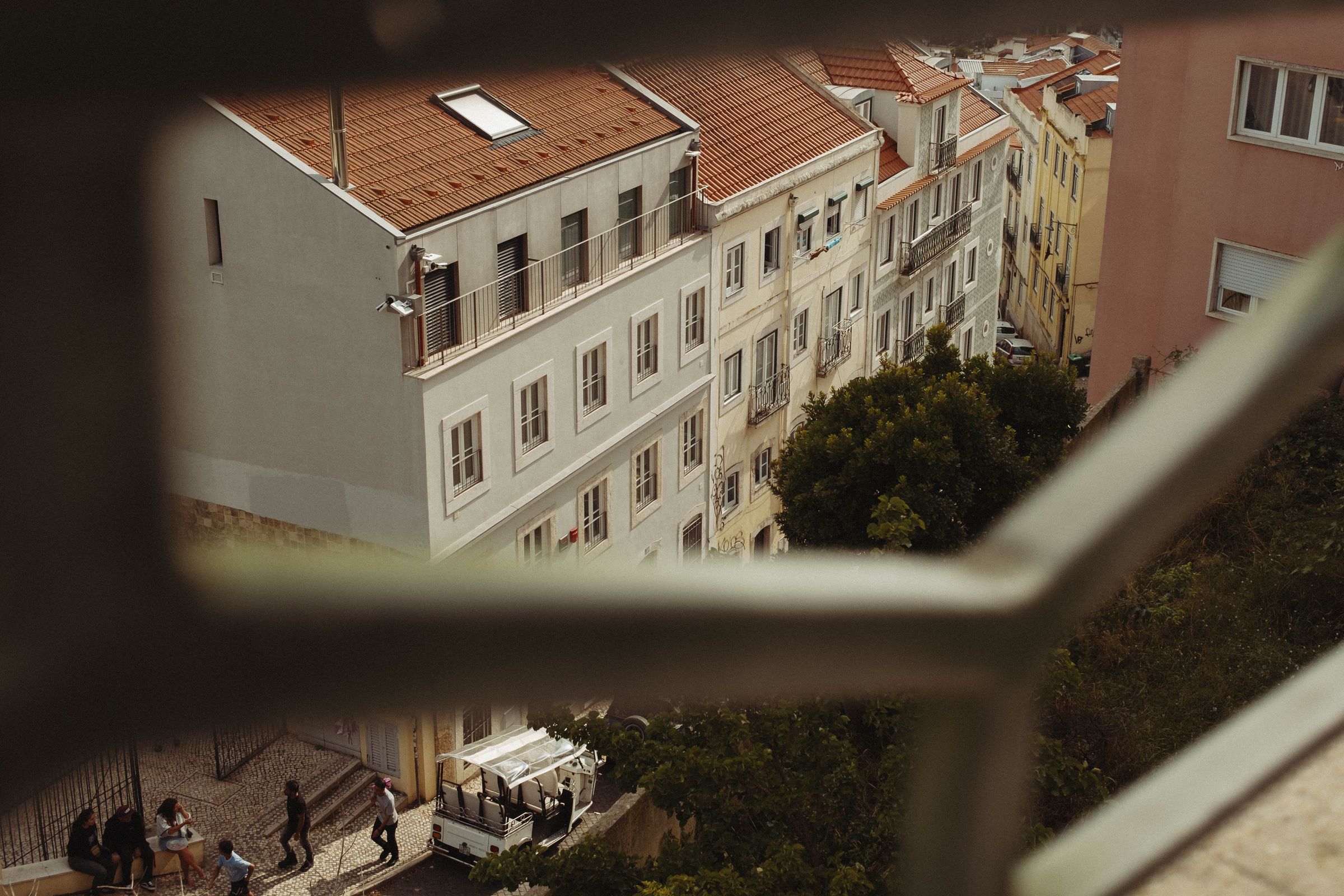 A view through a stone window opening frames Lisbon’s pastel-colored buildings with terracotta roofs clustered on a hillside. Below, pedestrians navigate a cobblestone street near a small outdoor café, while a green tree grows between the tightly packed structures.