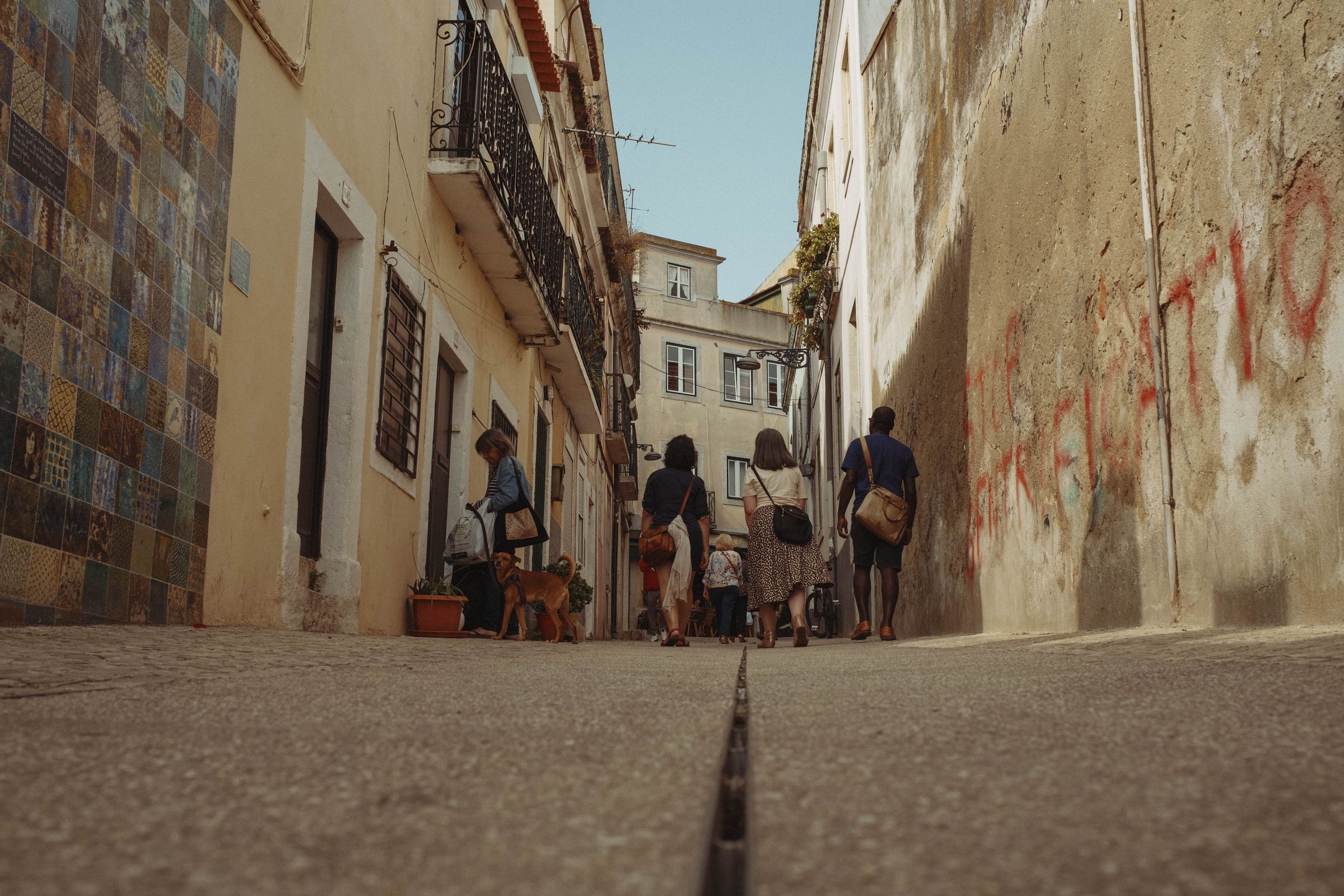 A group of pedestrians and a dog walk up a narrow, steep Lisbon street lined with weathered buildings, shot from ground level. Faded azulejo tiles decorate the left wall while peeling plaster marks the right, with wrought-iron balconies and terracotta rooftops visible above against a pale blue sky.