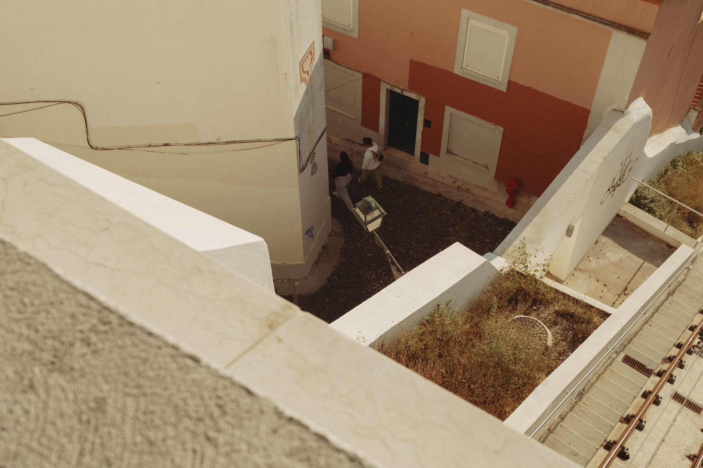 Looking down a series of rooftops with white painted walls, two figures stand in a small courtyard below. The geometric composition is framed by cream and coral-colored buildings with shuttered windows, their angles creating a striking bird’s-eye view of the narrow urban space.