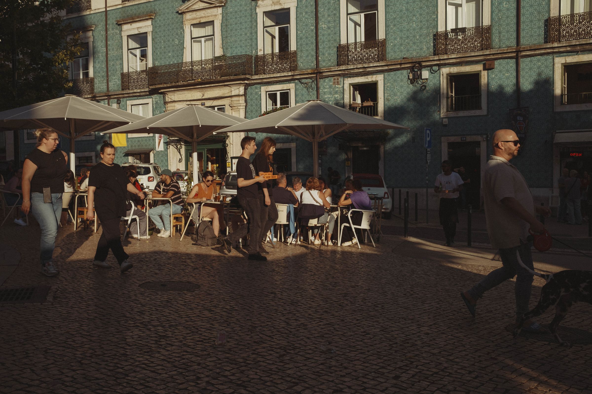 An outdoor café buzzes with activity in golden hour light, with servers carrying trays between tables of diners seated under tan umbrellas. The scene unfolds against a turquoise azulejo-tiled building with ornate iron balconies, while pedestrians cross the cobblestone square and dramatic shadows stretch across the pavement.