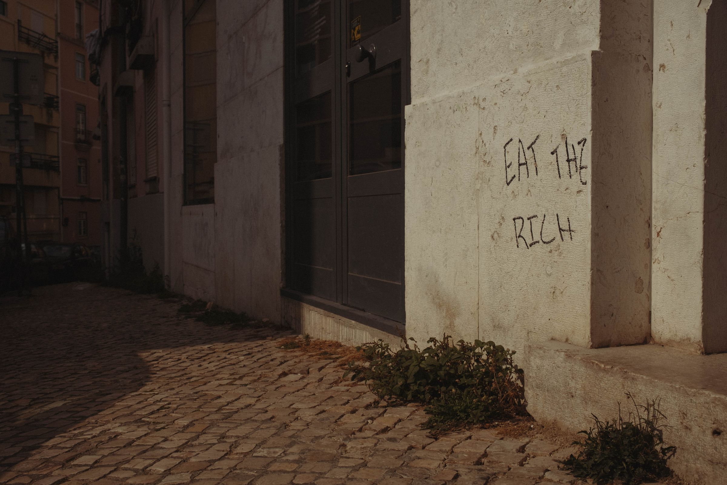Graffiti reading 'EAT THE RICH' is scratched into a pale stucco wall on a quiet cobblestone street corner, with weeds growing at its base. The stark message appears beside shuttered storefronts in the dim evening light, the narrow alley receding into shadow behind.