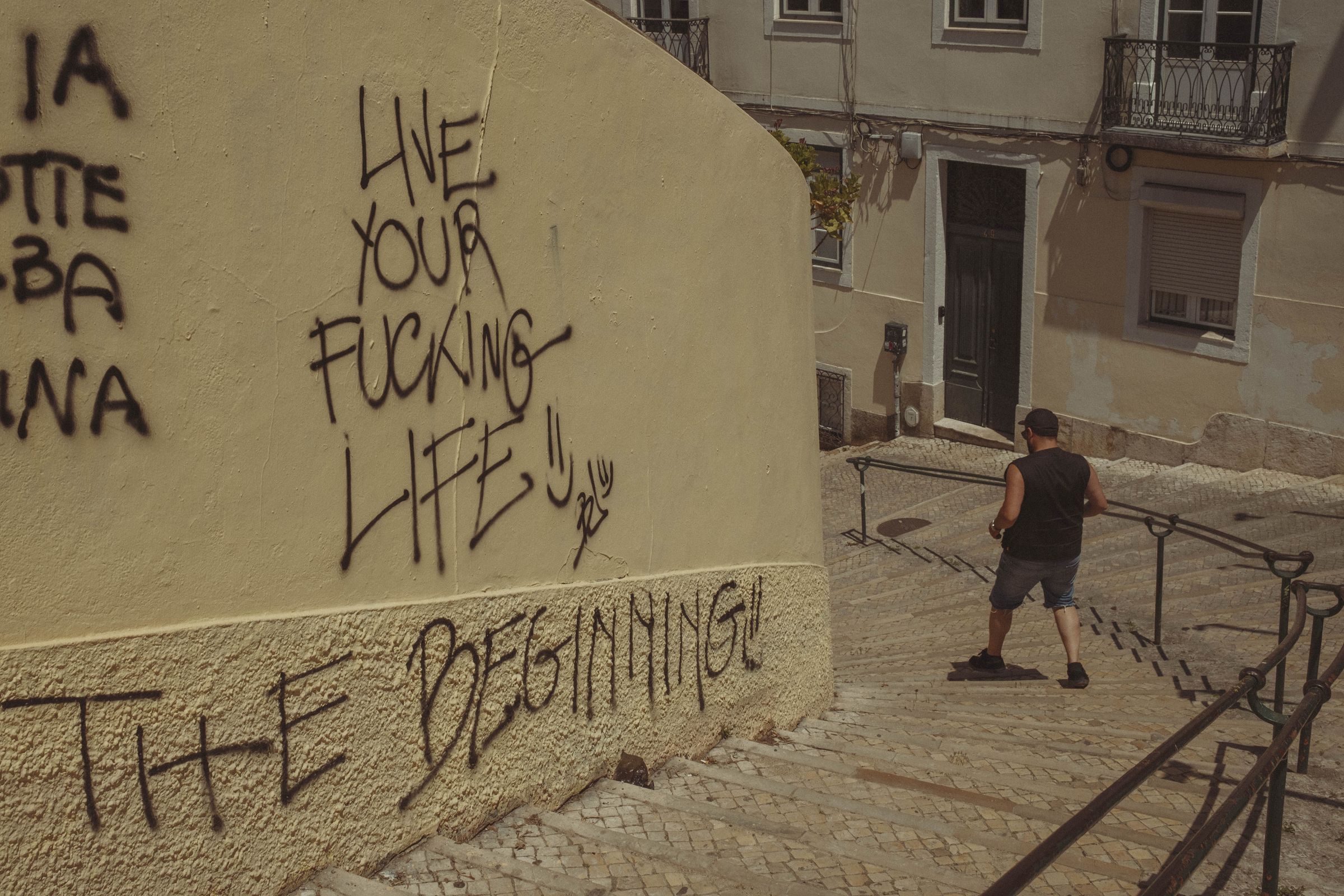 Large black graffiti reading 'LIVE YOUR FUCKING LIFE' and 'THIS IS BEGINNING!' covers a cream-colored wall on a sloped street, while a tourist in shorts descends the steps past the message. The defiant street art dominates the corner building above iron railings, contrasting with the quiet residential buildings beyond.
