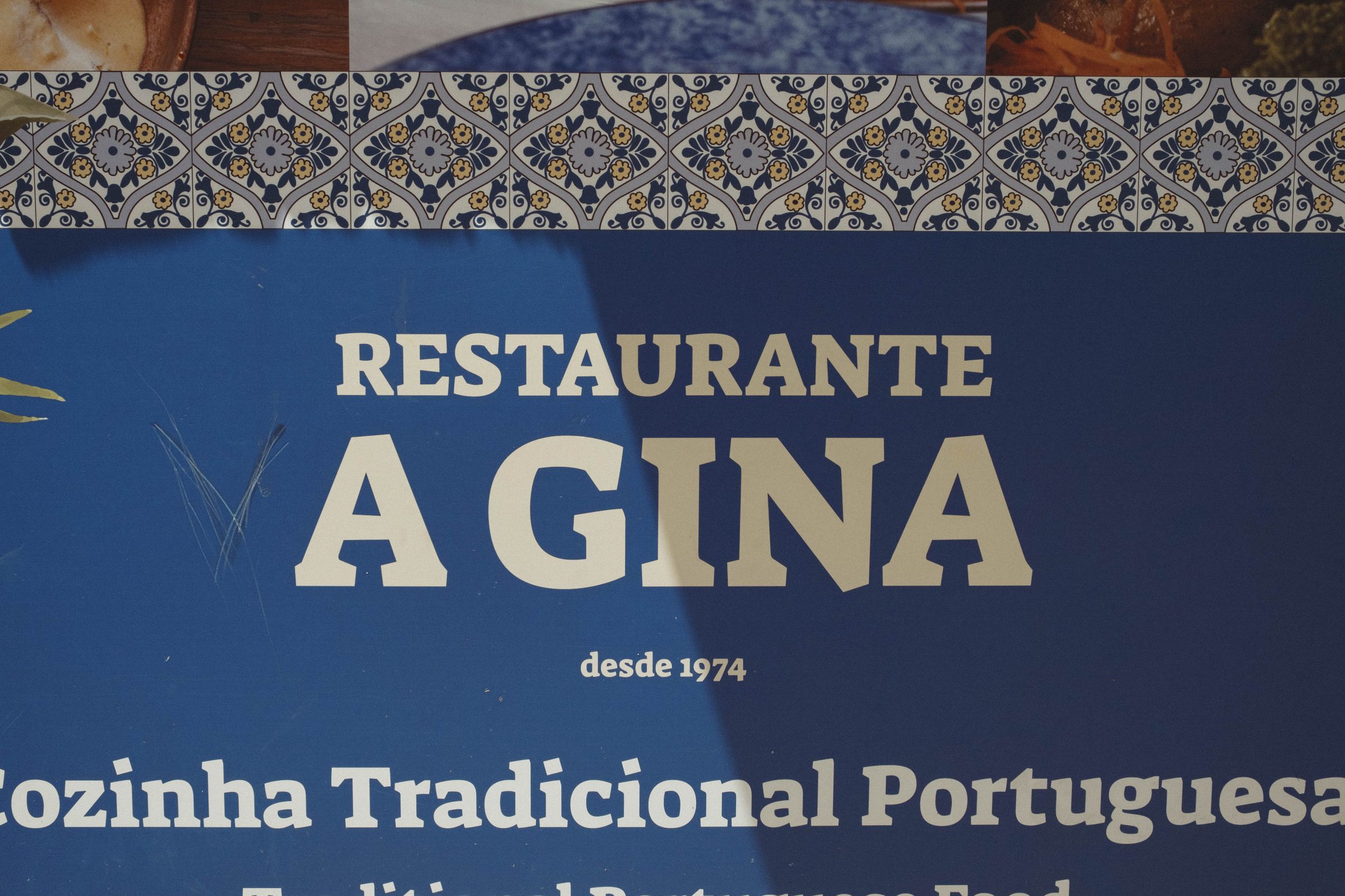 A weathered blue restaurant sign reading 'RESTAURANTE A GINA desde 1974' sits below a decorative border of traditional Portuguese azulejo tiles in blue and gold. The letter 'V' is etched onto the sign to spell 'VAGINA.' The scratched and faded signage advertises traditional Portuguese cuisine, showing decades of use since its 1974 founding.
