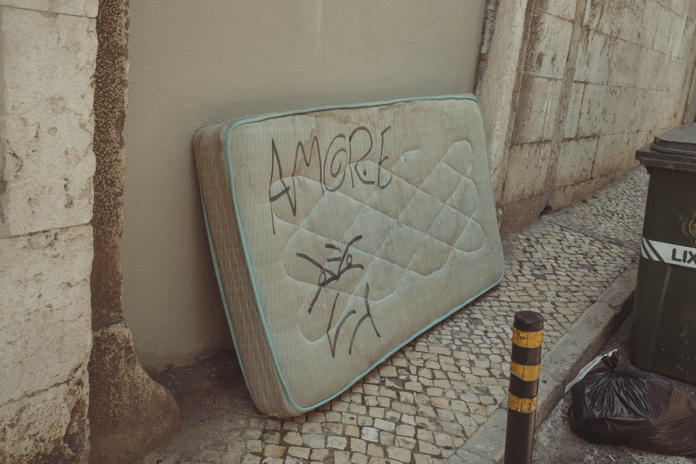 A discarded stained mattress with turquoise piping leans against a weathered wall, graffiti reading 'AMORE' and crude drawings marked across its quilted surface. The abandoned item rests on cobblestones beside trash bins and a bollard.