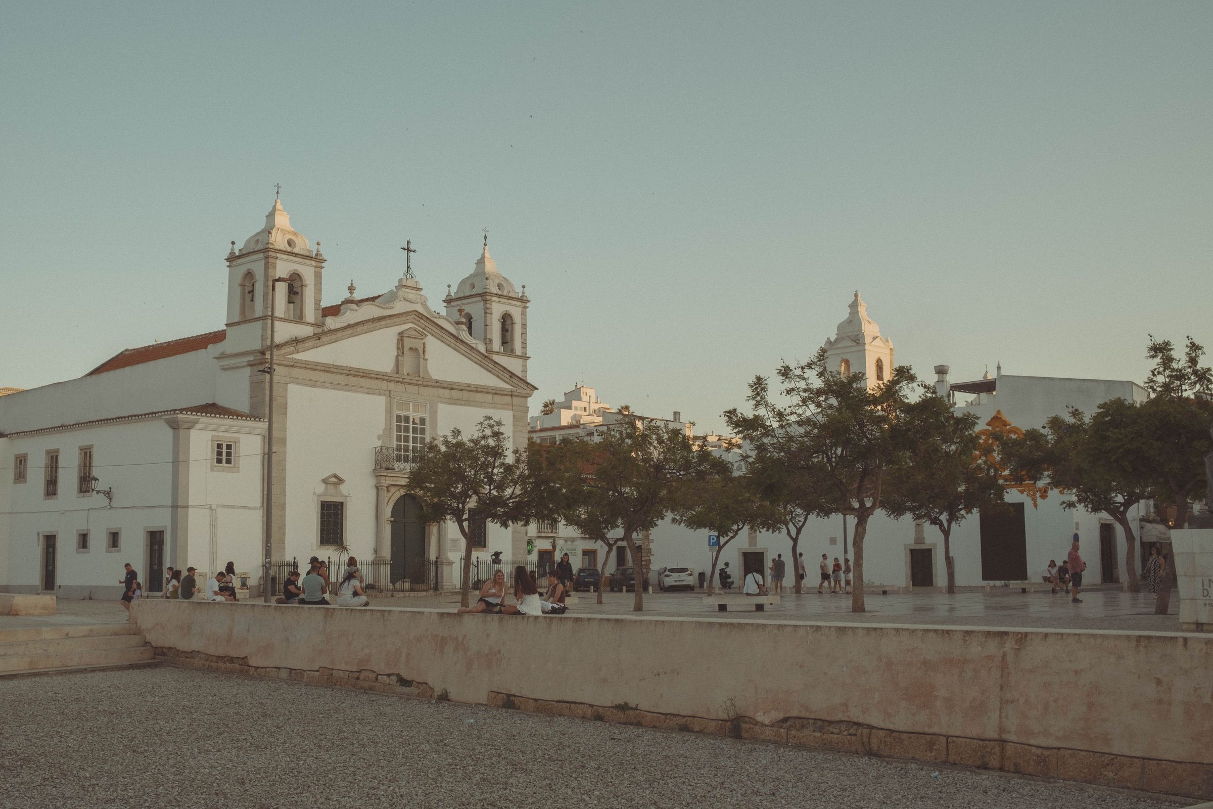 A whitewashed church with twin bell towers and terracotta roof anchors a plaza lined with trees, where people gather in the soft evening light. The baroque facade overlooks the square where locals and visitors sit on stone walls, with other church domes visible in the distance against a hazy sky.