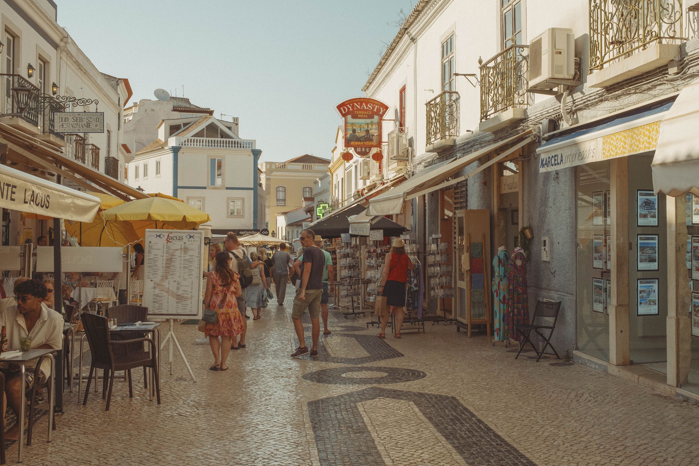 A narrow pedestrian street lined with shops and cafés draws tourists past souvenir stands and restaurant awnings in the old town. Whitewashed buildings with ornate iron balconies frame the cobblestone lane, where visitors browse postcards and outdoor dining tables fill the warm afternoon air.