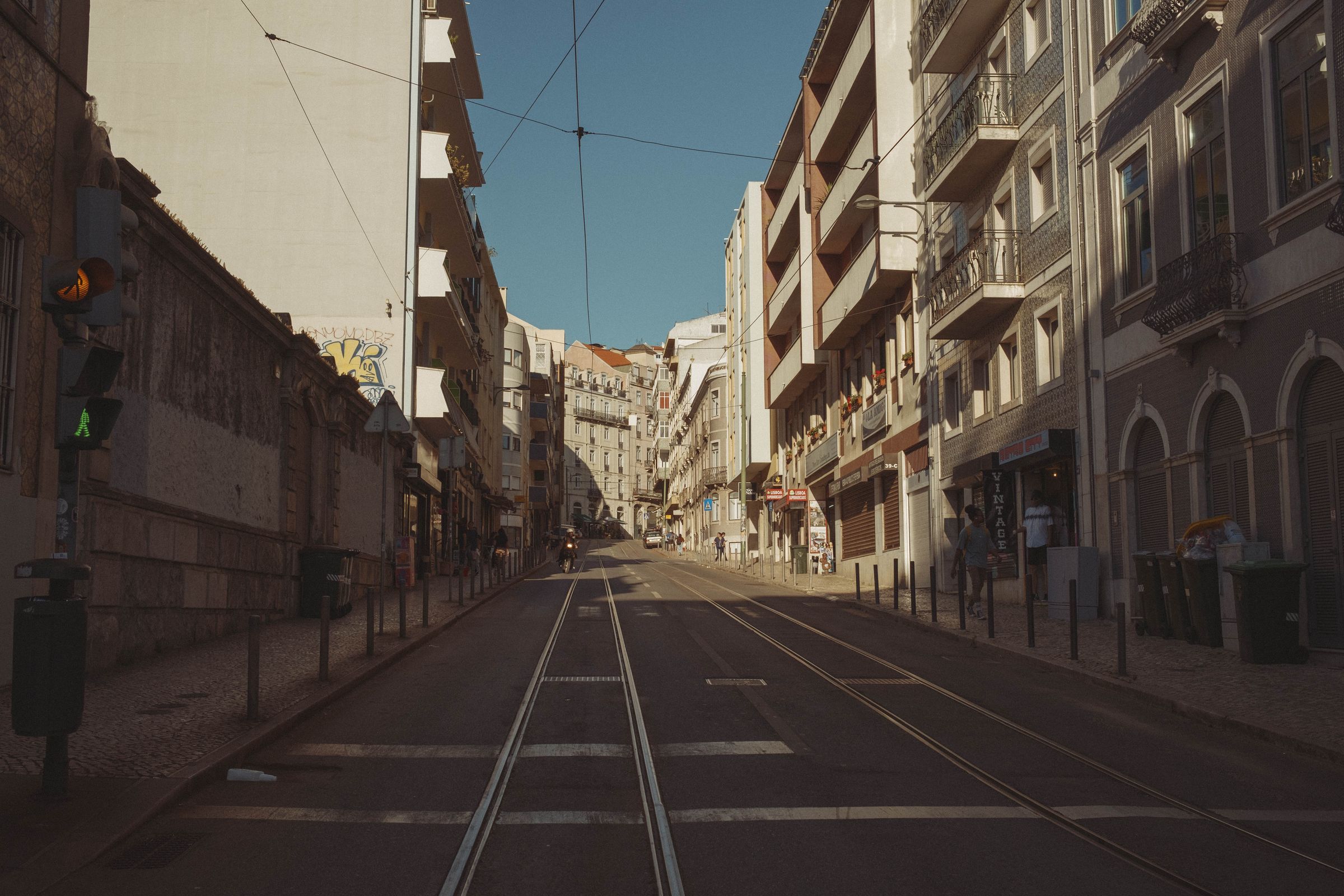 Tram tracks run up an empty Lisbon street at dawn, flanked by ornate apartment buildings with decorative facades and wrought-iron balconies. Overhead wires crisscross the clear blue sky as golden light catches the architecture, while shuttered storefronts line the quiet cobblestoned road.