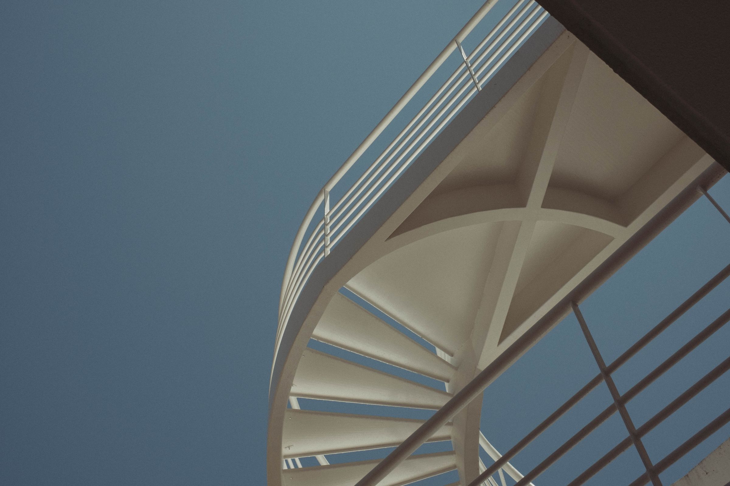 A white modernist pedestrian bridge curves gracefully across a clear blue sky, its geometric understructure creating bold angular shadows. The clean concrete arch with parallel suspension cables creates a striking composition of light and form against the cloudless afternoon sky.Claude is AI and can make mistakes. Please double-check responses.