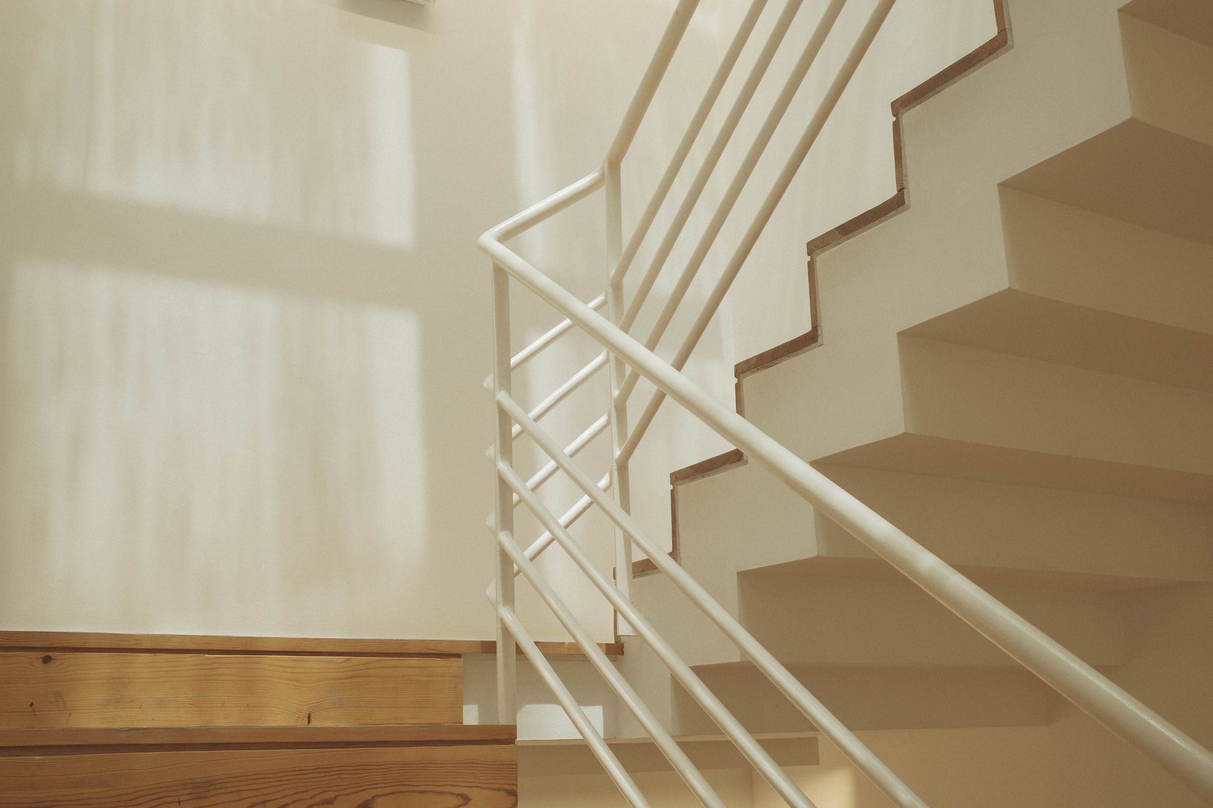 A minimalist staircase with blonde wood treads ascends along cream-colored walls, its white metal railing creating clean geometric lines. Natural light filters through, casting soft shadows across the pristine surfaces of the modern interior.