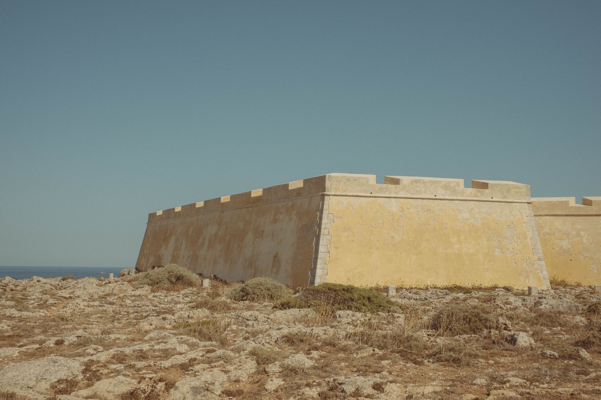 Massive stone fortification walls angle sharply upward from sparse, rocky terrain, their weathered surfaces showing variations from golden to pale cream. The geometric defensive structure meets the Atlantic horizon, its crenellated ramparts silhouetted against a clear blue sky.