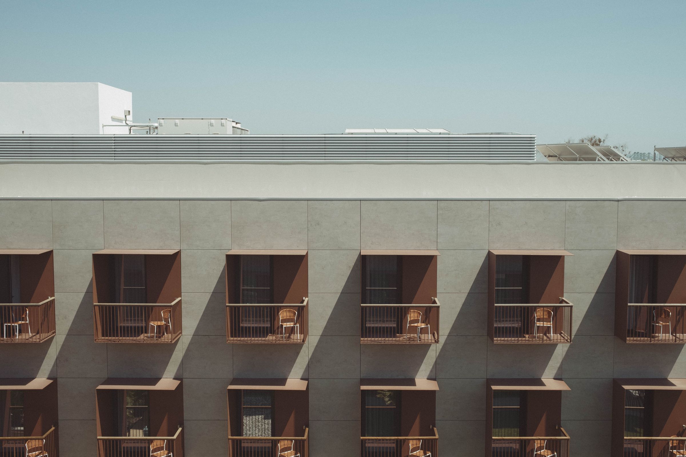 A minimalist hotel facade features rows of identical balconies with wooden railings and wicker chairs, their geometric pattern creating sharp shadows in bright sunlight. The pale gray concrete structure contrasts with the clear blue sky, while white buildings are visible in the distance along the roofline.