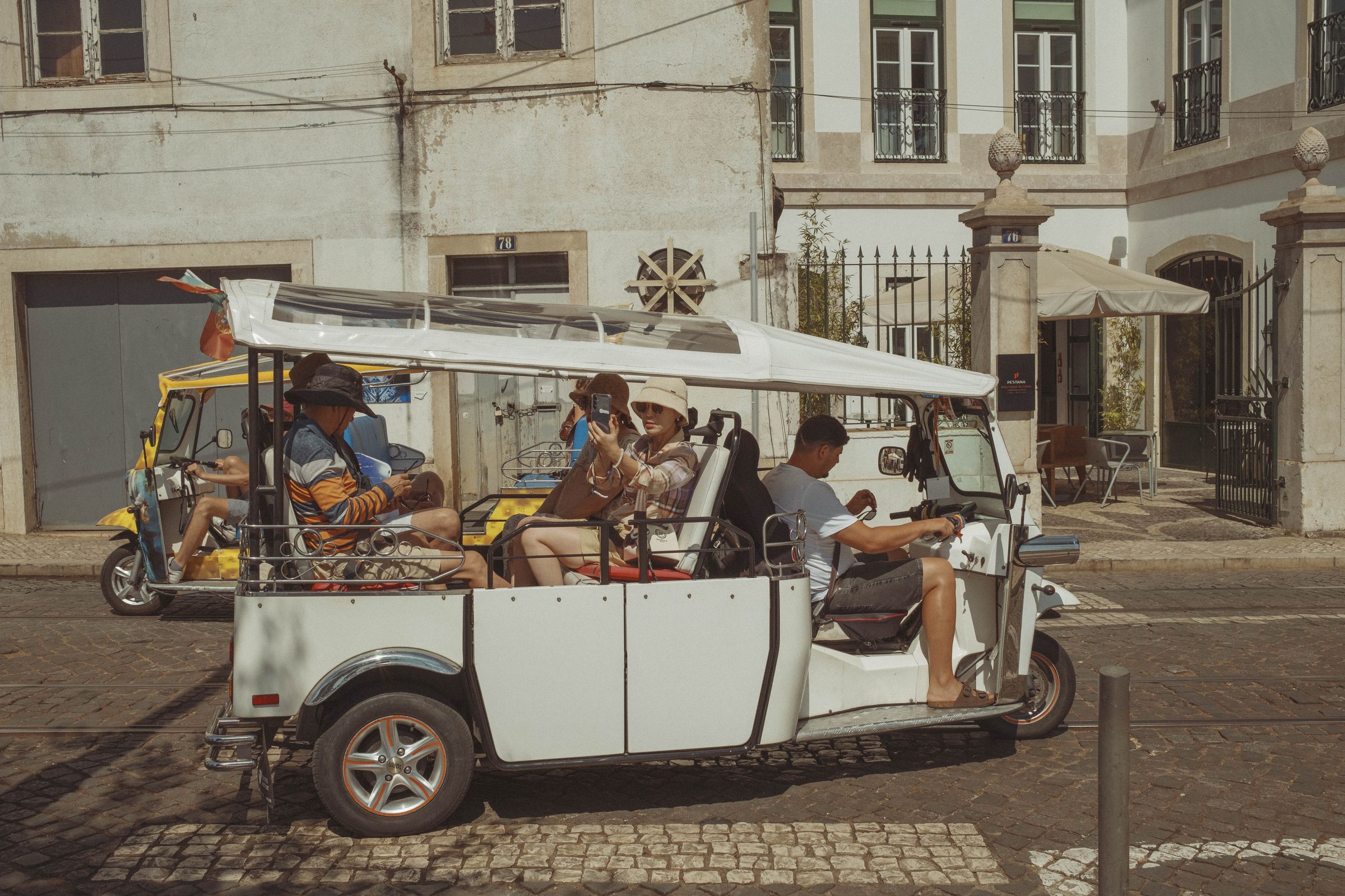 Tourists sitting in a white golf cart through a historic neighborhood, with passengers snapping photos as they sit near weathered buildings and an iron-gated entrance. The open-air vehicle is next to stained facades and cobblestone streets.