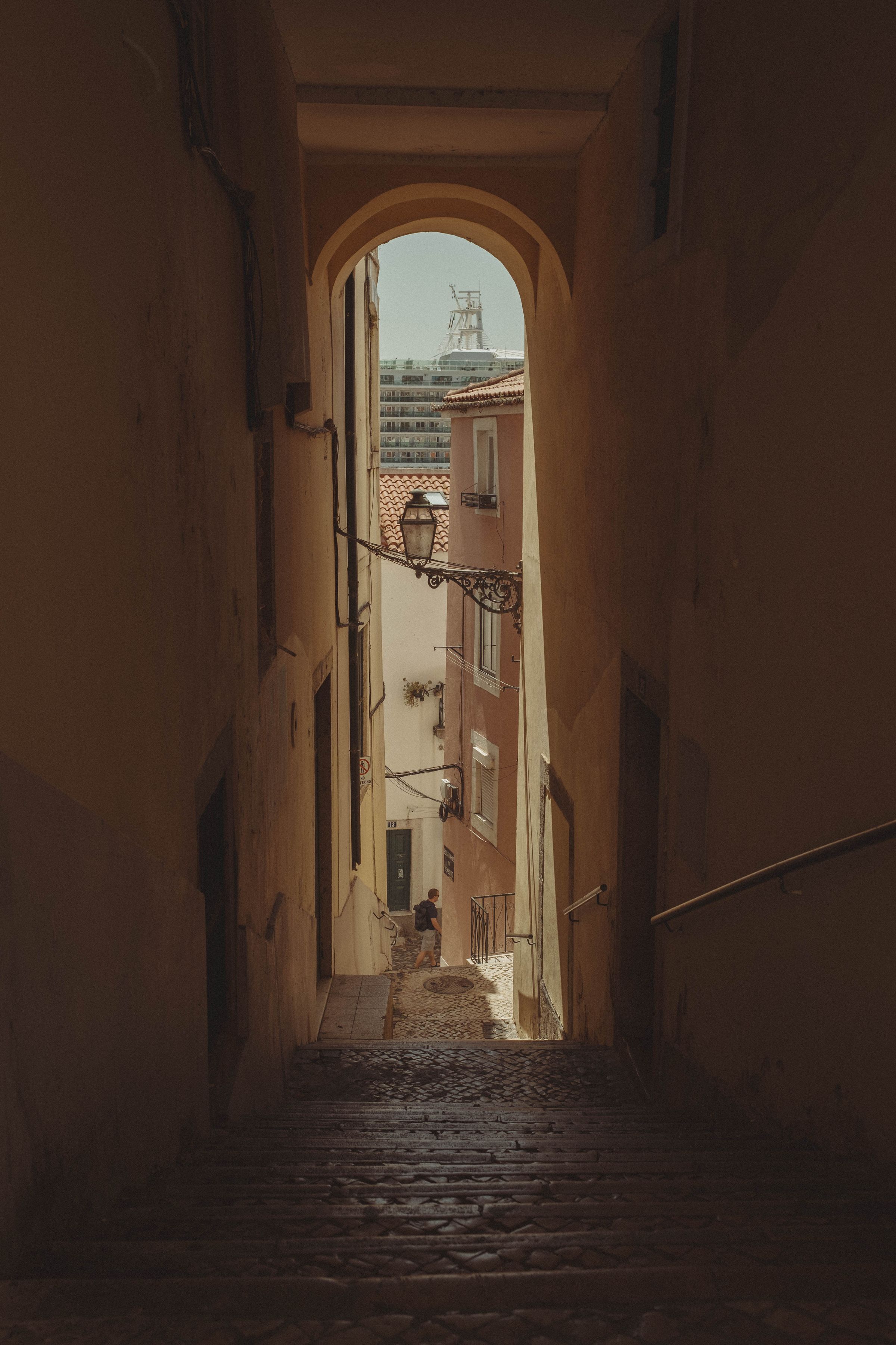 A stone archway frames a narrow cobbled lane descending between salmon-pink buildings, with an ornate iron lantern hanging against the aged walls. A lone figure sits in the shadowed alleyway while cruise ship can be seen in the distance, the worn steps leading down toward brighter light below.