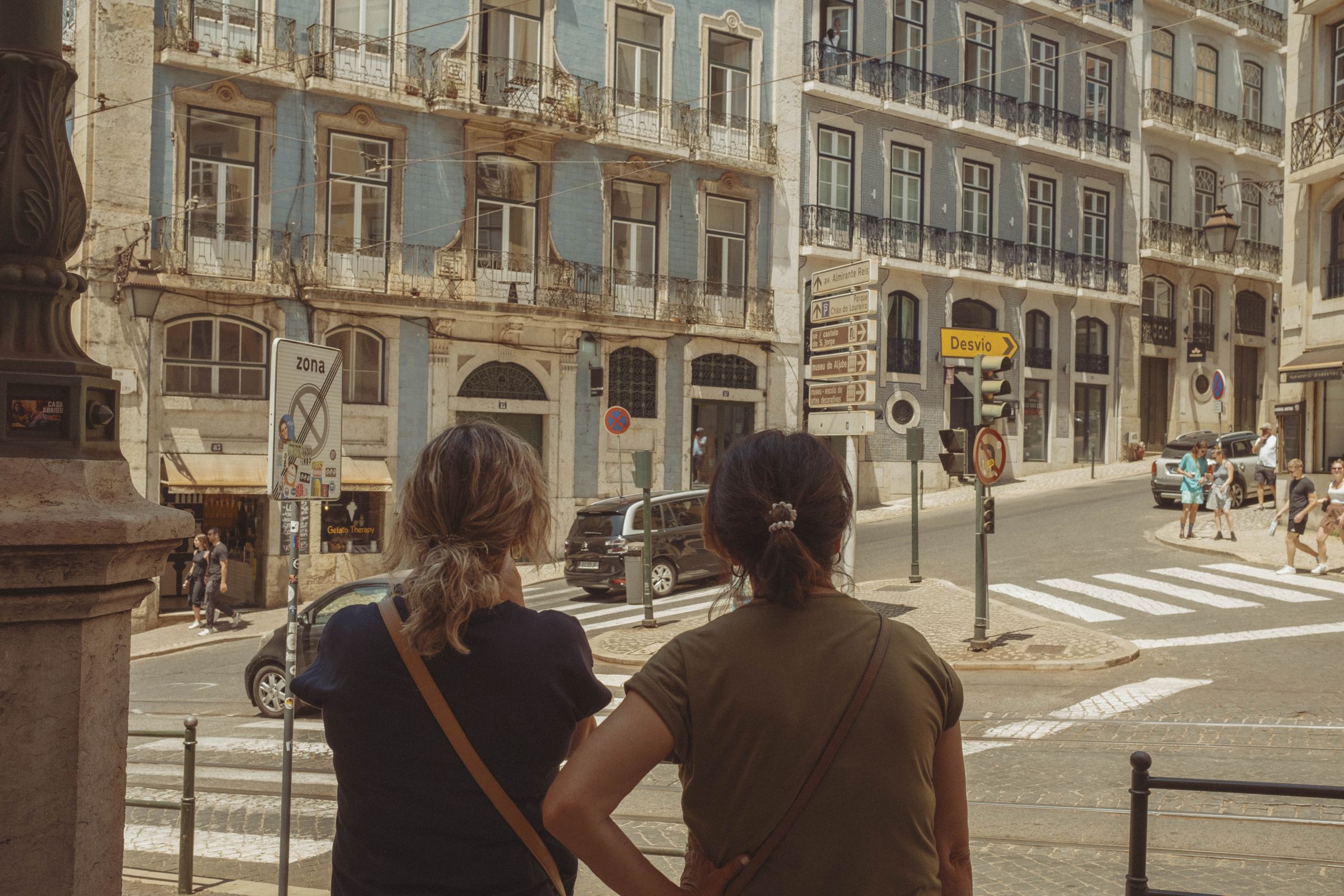Two women stand at an intersection photographing weathered apartment buildings with faded blue and cream facades and ornate iron balconies. Pedestrians cross the zebra crossing beyond them while cars navigate the busy junction, the worn architectural grandeur contrasting with the everyday bustle of the downtown street.