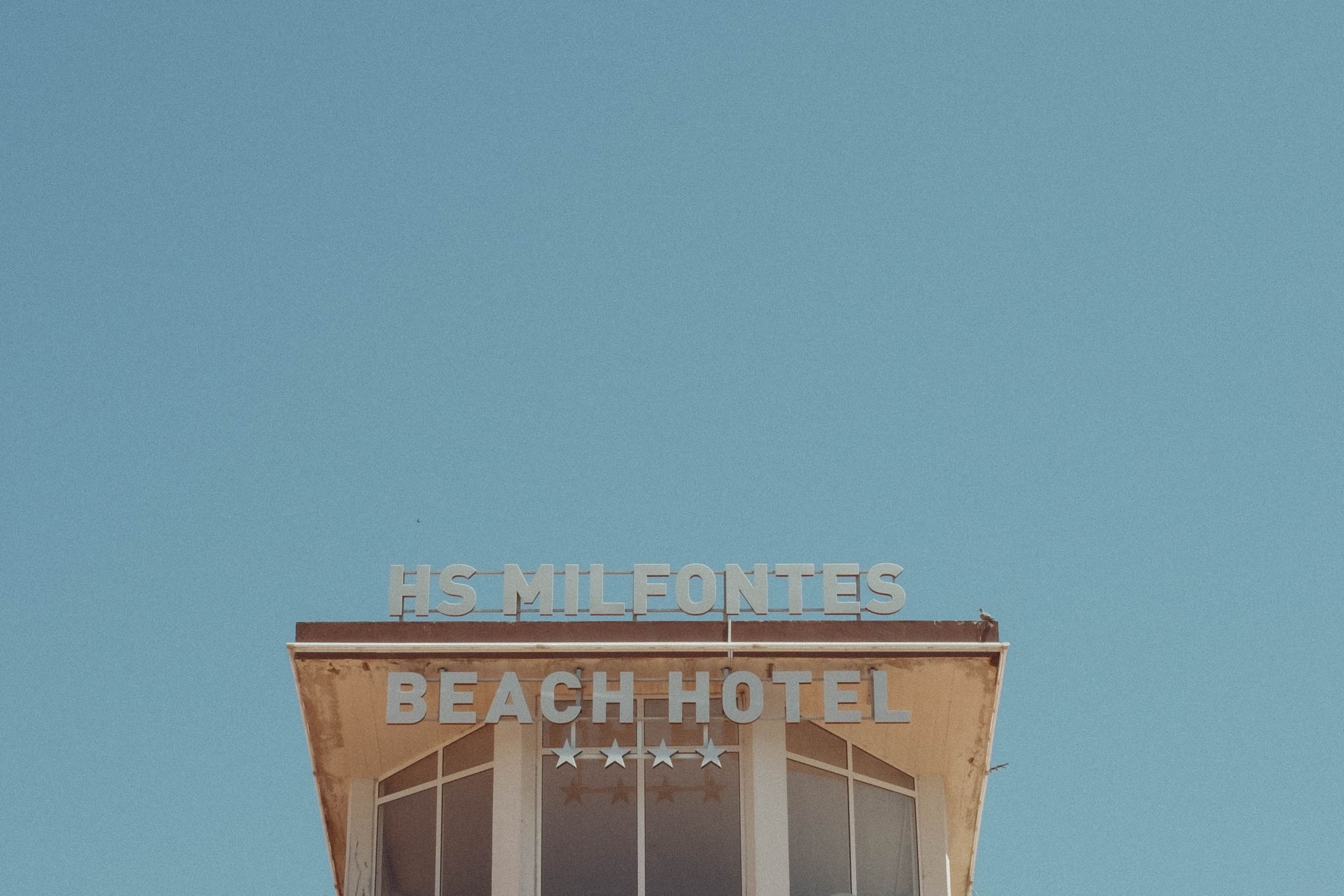 The weathered rooftop sign of HS Milfontes Beach Hotel displays faded lettering and four stars against a pale blue sky. The beige facade shows signs of age, with the white block letters standing in stark contrast to the worn paint below.